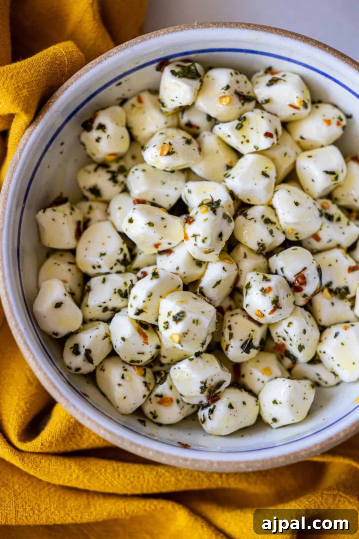 A delightful bowl brimming with marinated mozzarella cheese balls, each a small sphere of creamy perfection seasoned with a medley of aromatic herbs and savory spices, resting on a soft yellow cloth. An irresistible Mozzarella Appetizer ready to be savored.