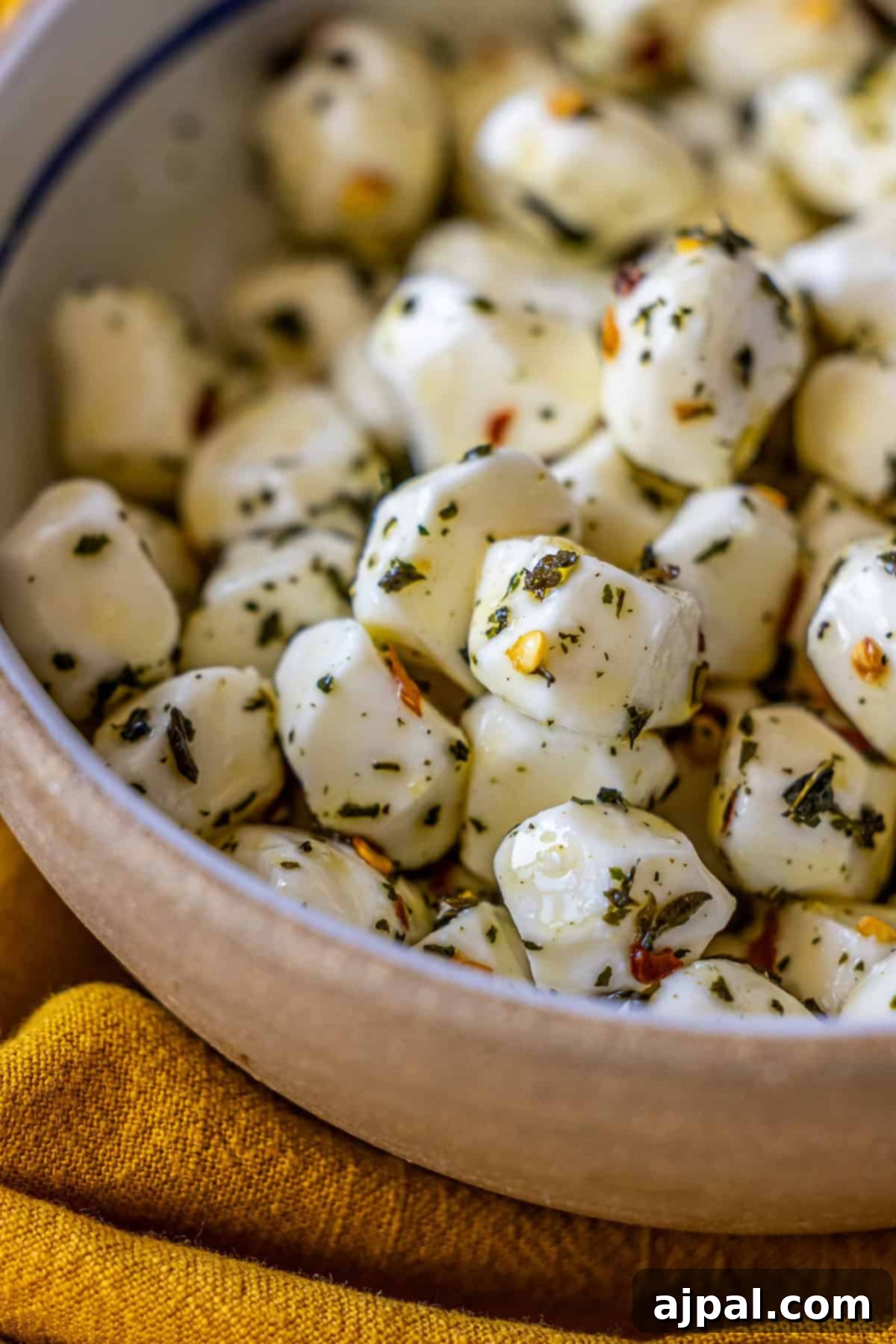 A vibrant bowl filled with marinated mozzarella cheese balls, generously seasoned with a rich blend of herbs and red pepper flakes, all nestled on a warm yellow cloth. This simple yet sophisticated appetizer is ready to delight your guests.