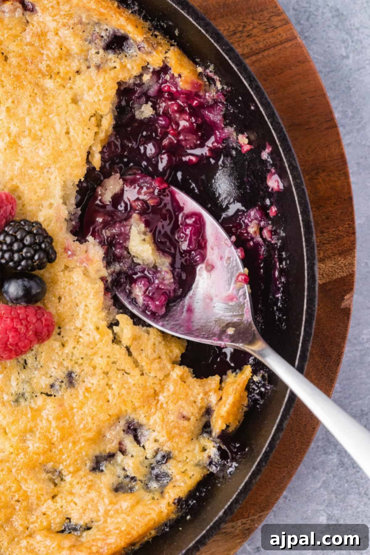 A close-up of a Smoked Berry Cobbler in a skillet, with a spoon scooping out some filling and berries on top as garnish.