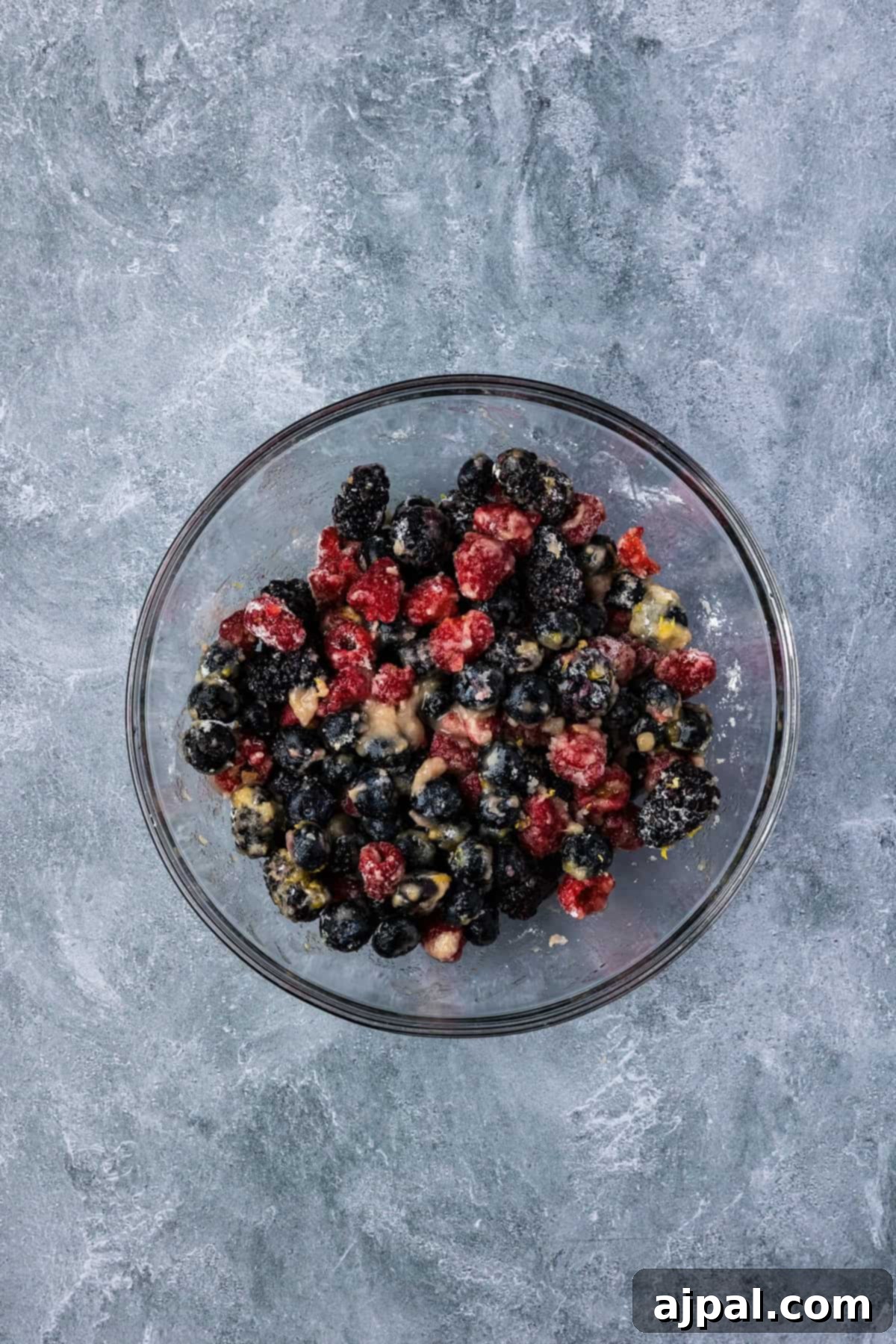 A glass bowl filled with a mixture of blackberries, blueberries, and raspberries, coated with sugar, perfect for creating a delicious Smoked Berry Cobbler, sits on a gray textured surface.
