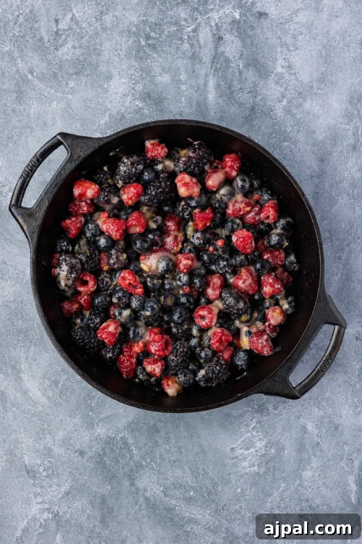 A cast iron skillet filled with a mixture of raspberries, blackberries, and blueberries creates a vibrant Smoked Berry Cobbler on a gray textured surface.