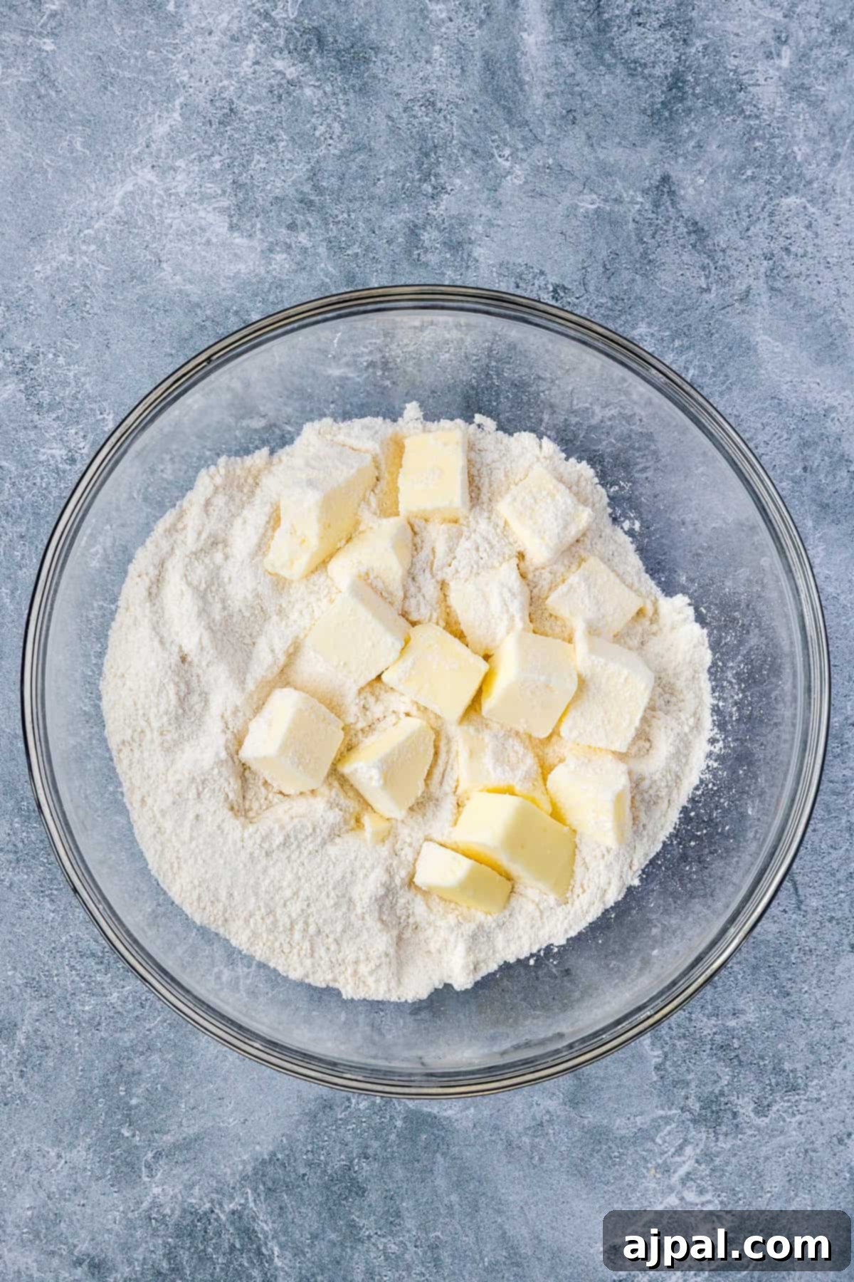 A glass bowl filled with flour and cubes of butter, ready to be transformed into a delicious Smoked Berry Cobbler, sits on a gray textured surface.