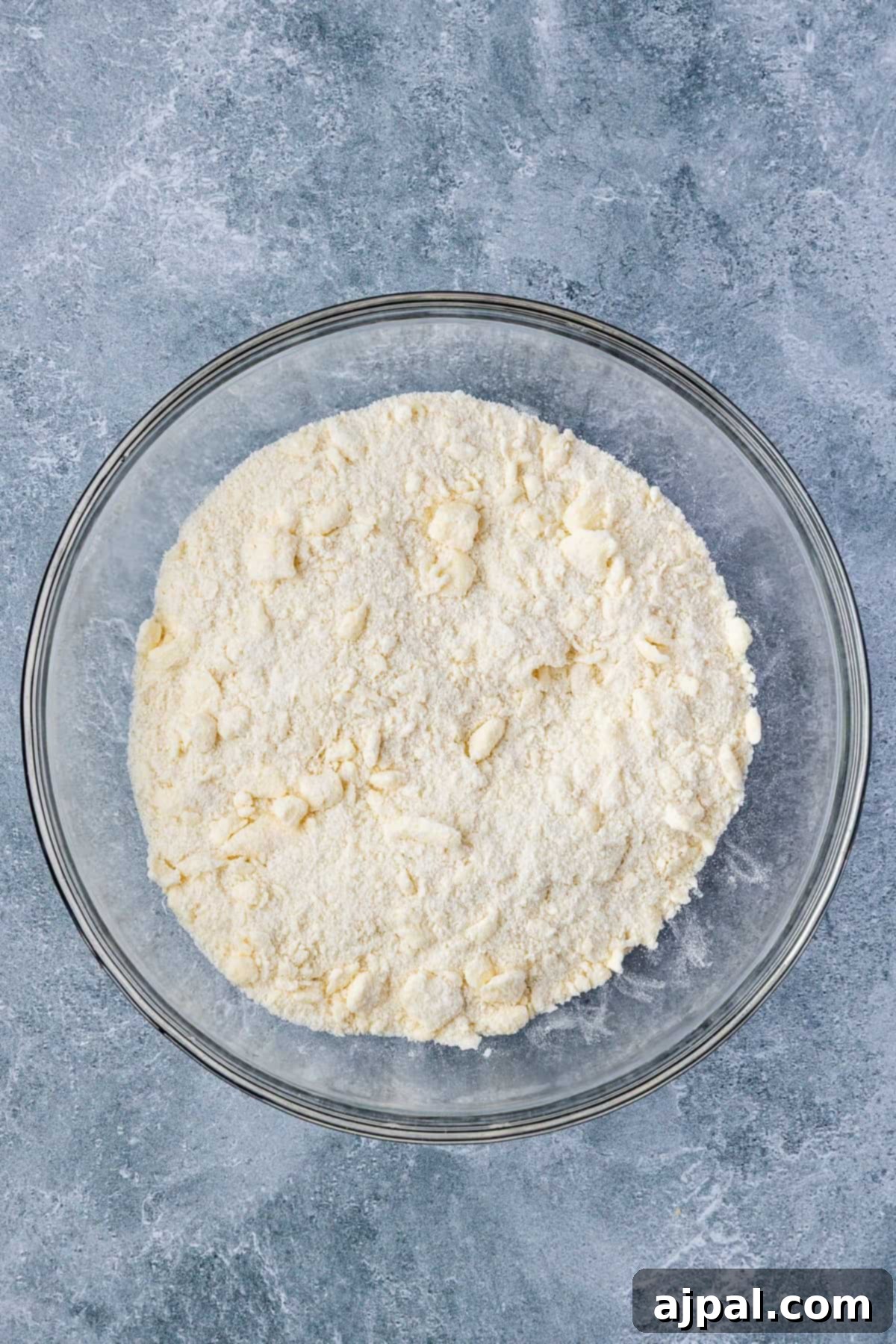 A glass bowl filled with flour and small pieces of butter sits on a gray textured surface, ready to become the perfect base for a delicious Smoked Berry Cobbler.