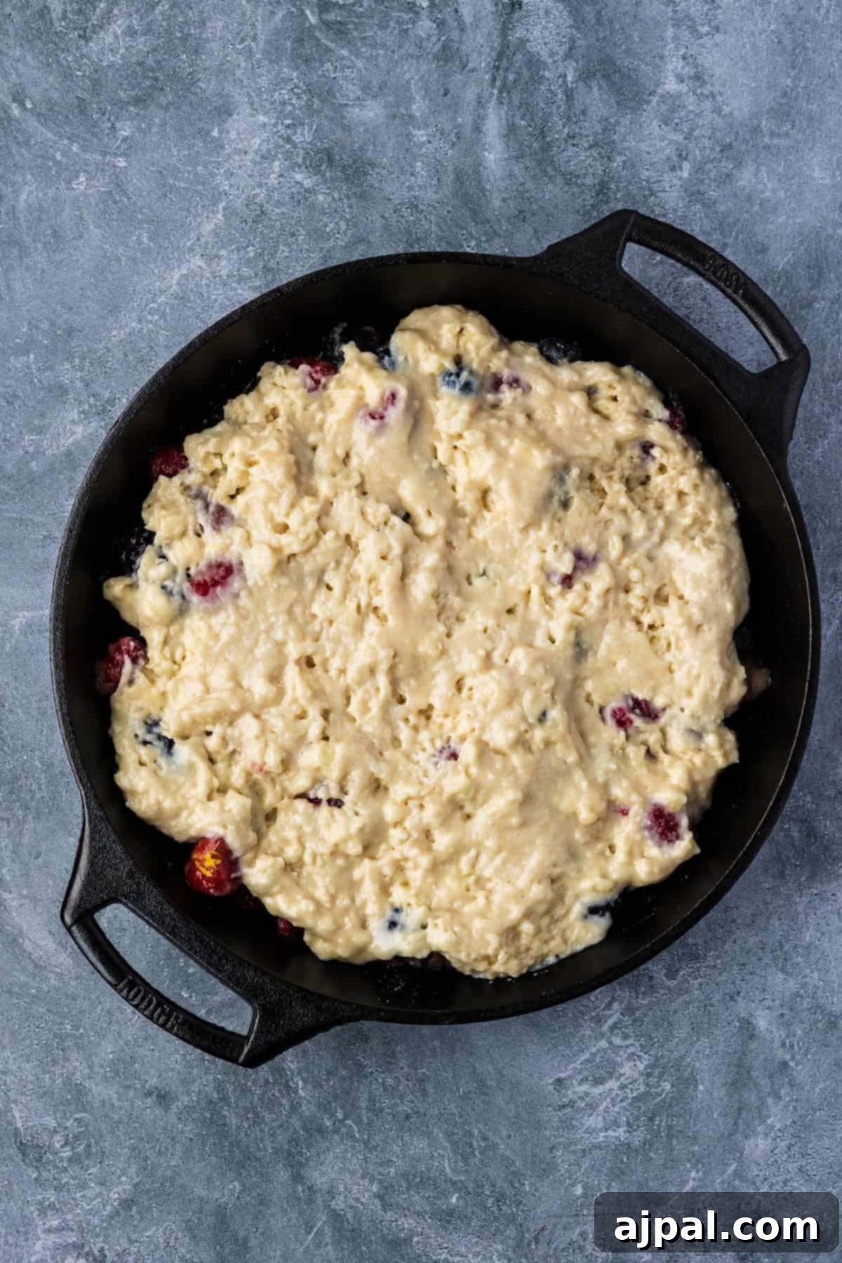 A cast iron skillet filled with unbaked Smoked Berry Cobbler dough sits on a gray textured surface.
