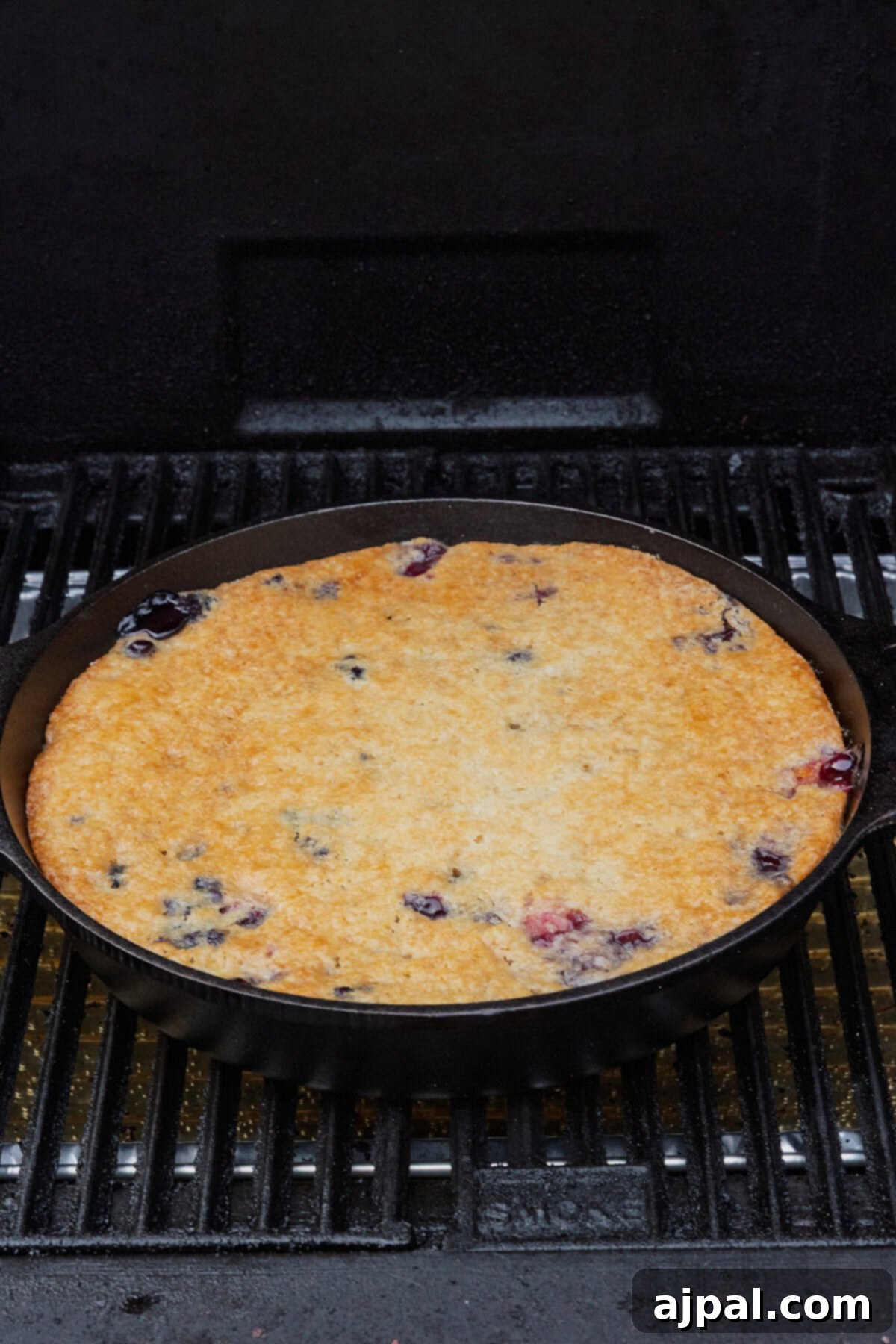 A cast iron skillet with a Smoked Berry Cobbler sits on a grill grate, its golden crust bubbling with juicy blueberries.