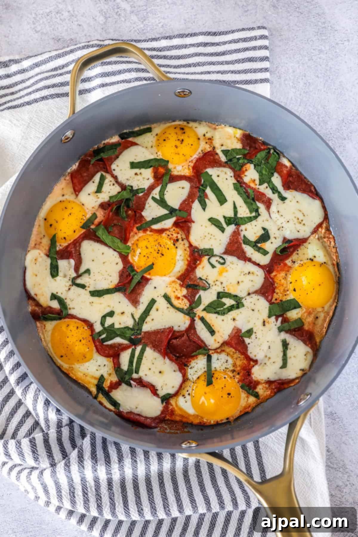 A skillet filled with baked eggs, melted mozzarella cheese, pepperoni slices, and fresh basil creates a delicious keto breakfast pizza, resting on a striped kitchen towel.