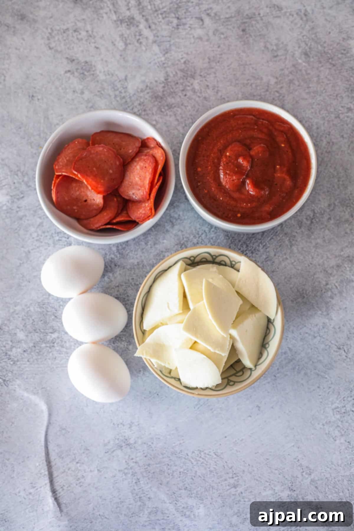 Three fresh eggs, a bowl of thinly sliced pepperoni, a small bowl of rich red pizza sauce, and a generous heap of shredded mozzarella cheese are artfully arranged on a clean grey surface. These are all the vibrant, essential ingredients you need to create a truly delicious and satisfying keto breakfast pizza at home.