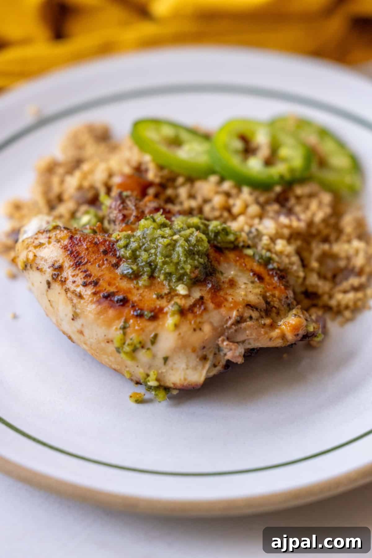 A perfectly grilled Zhoug chicken breast, exquisitely topped with a vibrant green sauce, accompanied by a side of fluffy couscous and delicate slices of jalapeños, all presented on a pristine white plate.