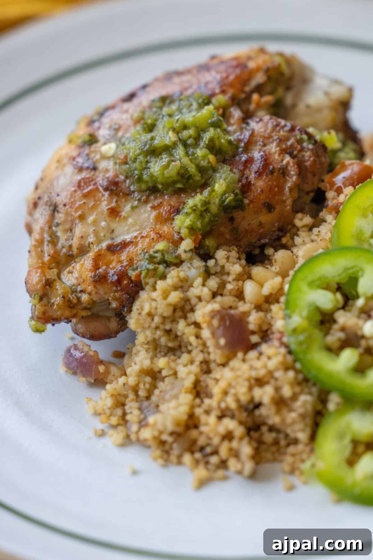Exquisitely grilled Zhoug Chicken thighs, shimmering with green sauce, beautifully arranged on a white plate next to seasoned couscous, fresh green beans, and thinly sliced jalapeño peppers.