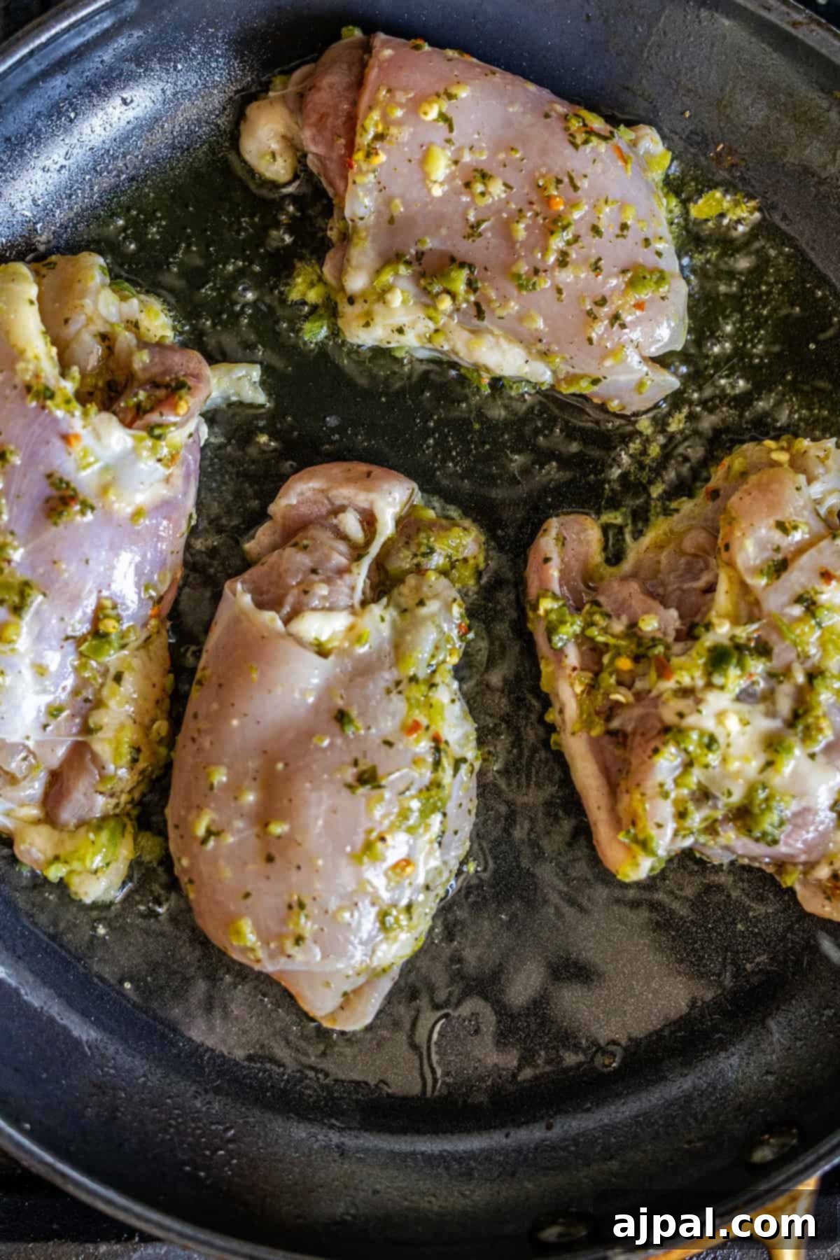 Raw, herbaceous Zhoug marinated chicken thighs sizzling vigorously in hot oil within a sleek black skillet, indicating the start of a flavorful searing process.