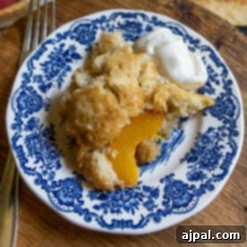 Tangy Sourdough Peach Cobbler 10 A perfectly portioned slice of sourdough peach cobbler, crowned with a dollop of fluffy whipped cream, rests on a charming blue and white floral-patterned plate, with a fork at its side on a rustic wooden surface.