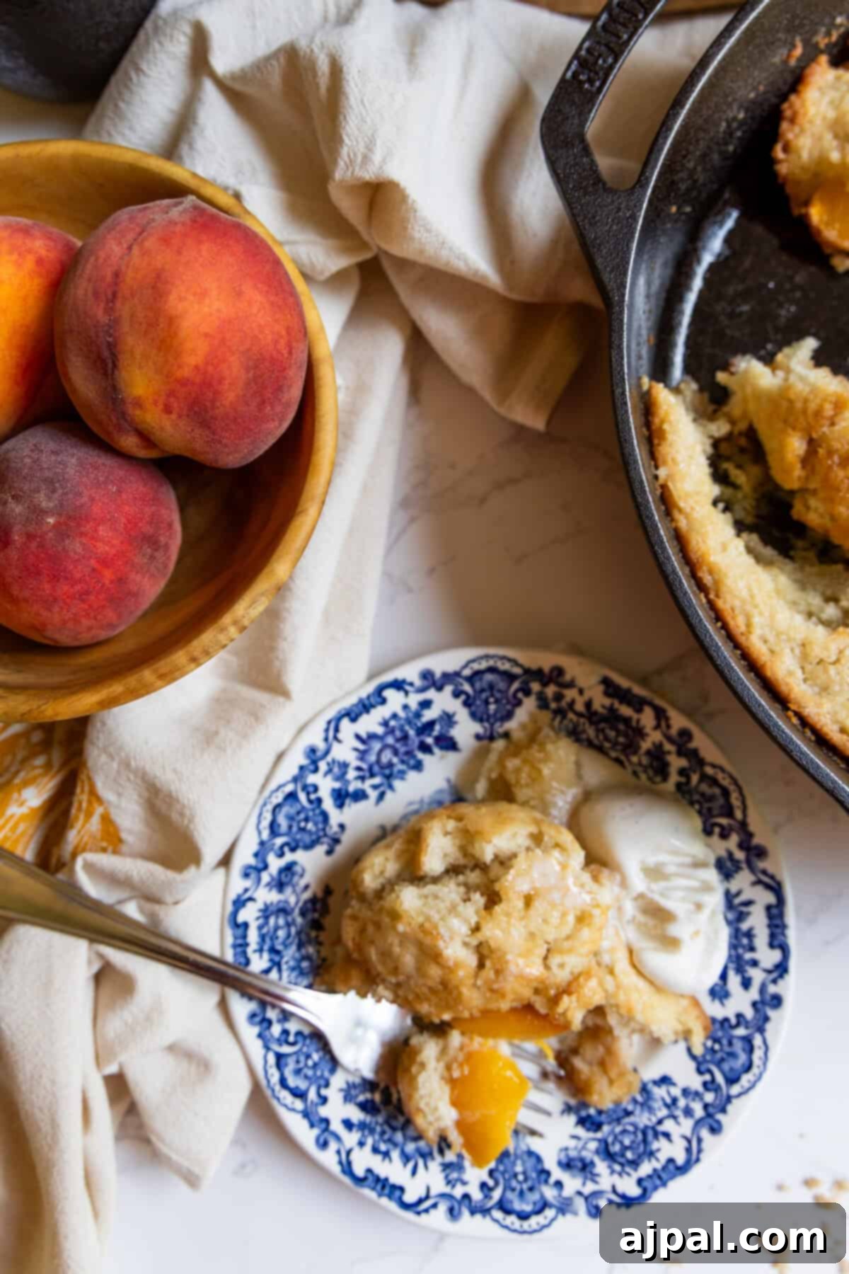 Tangy Sourdough Peach Cobbler 5 A beautiful presentation of sourdough peach cobbler: a single serving with ice cream on a blue and white plate, beside a cast iron skillet of fresh cobbler and a wooden bowl brimming with fresh peaches.
