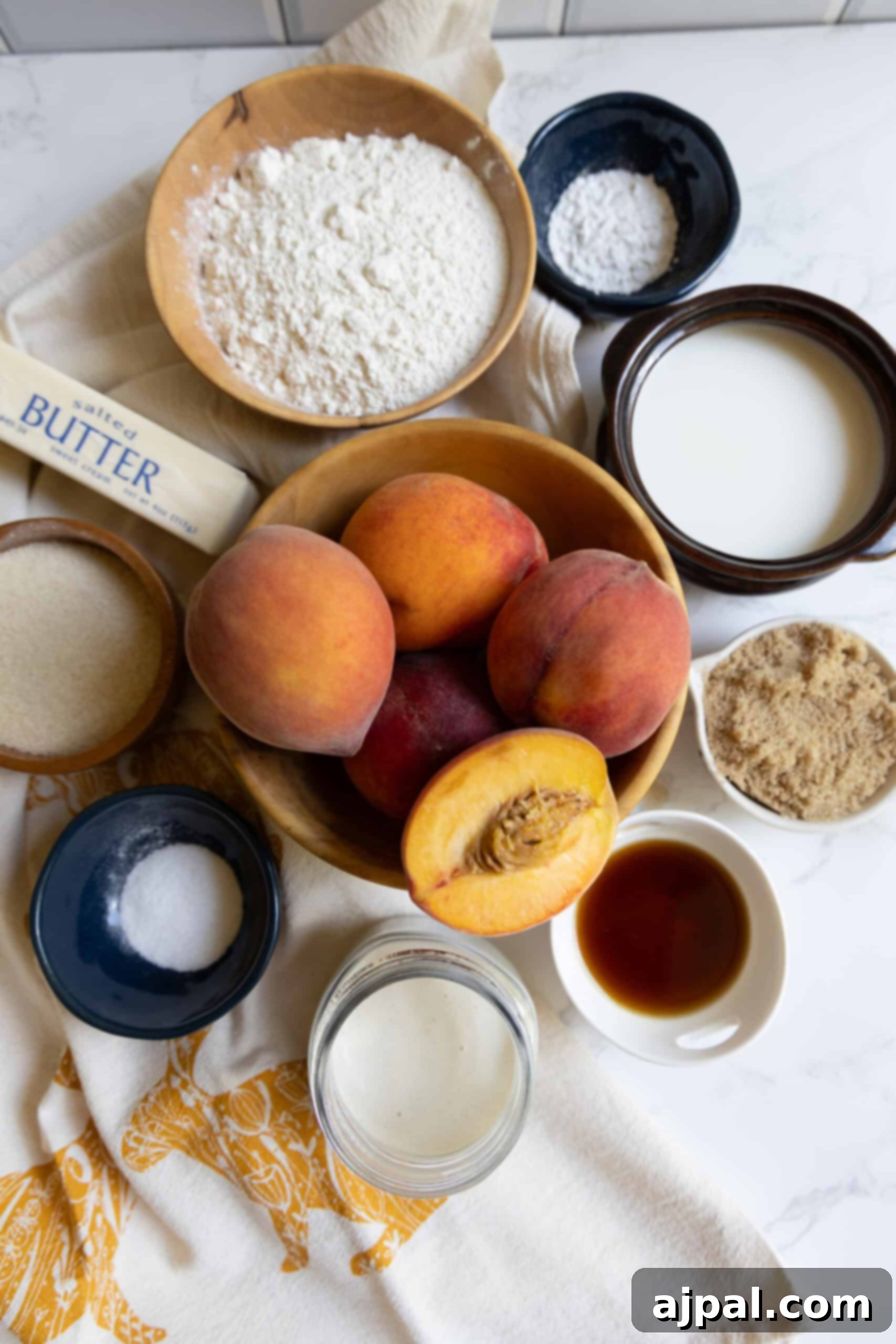 Tangy Sourdough Peach Cobbler 6 A flat lay photograph of all baking ingredients for sourdough peach cobbler, including fresh peaches, flour, granulated sugar, brown sugar, butter, milk, vanilla extract, baking powder, and salt, neatly arranged on a rustic kitchen towel.