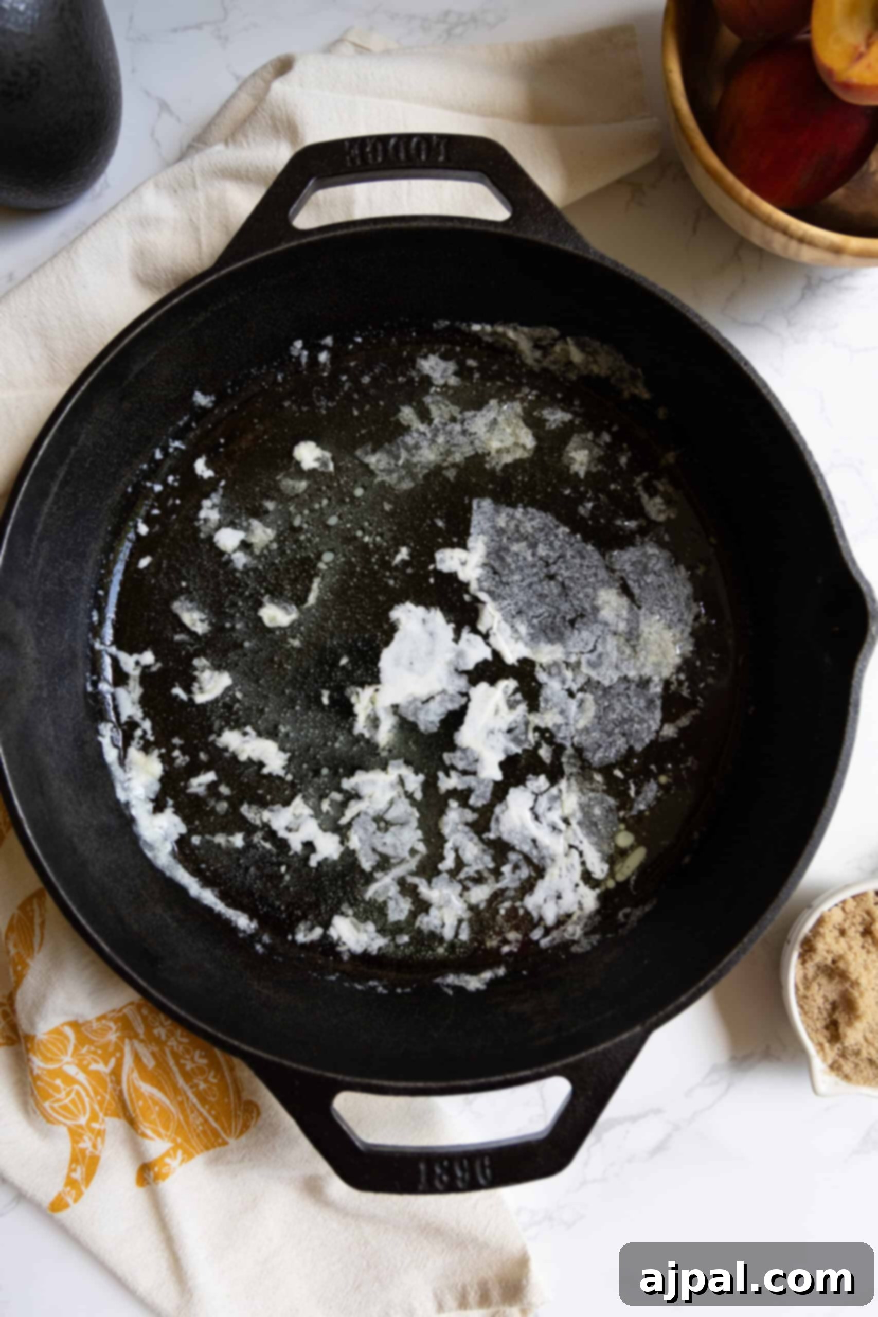 Tangy Sourdough Peach Cobbler 7 A cast iron skillet sits on a patterned cloth, containing partially melted butter, surrounded by a bowl of fresh peaches and a dish of brown sugar, setting the scene for baking.
