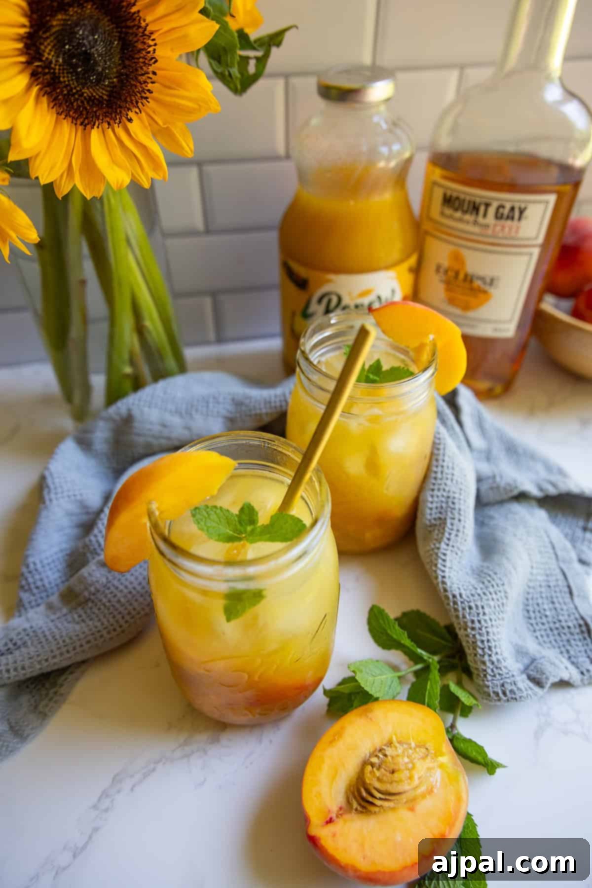 Two mason jars filled with a bourbon peach smash cocktail, garnished with peach slices and mint. A bottle of rum, peach juice, fresh peaches, and sunflowers are in the background.