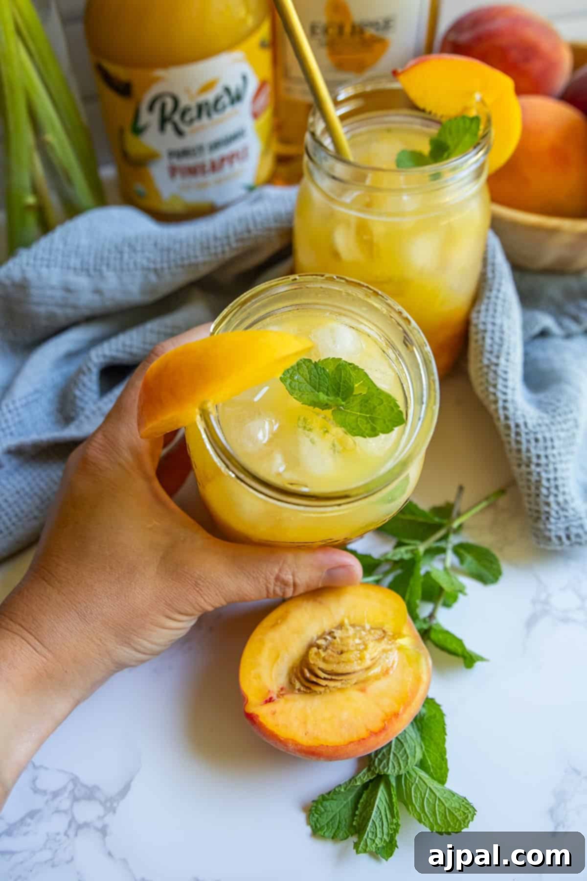 Hand holding a glass of bourbon peach smash cocktail garnished with a peach slice and mint; another glass, a bottle of juice, peaches, and mint are on a white surface in the background.