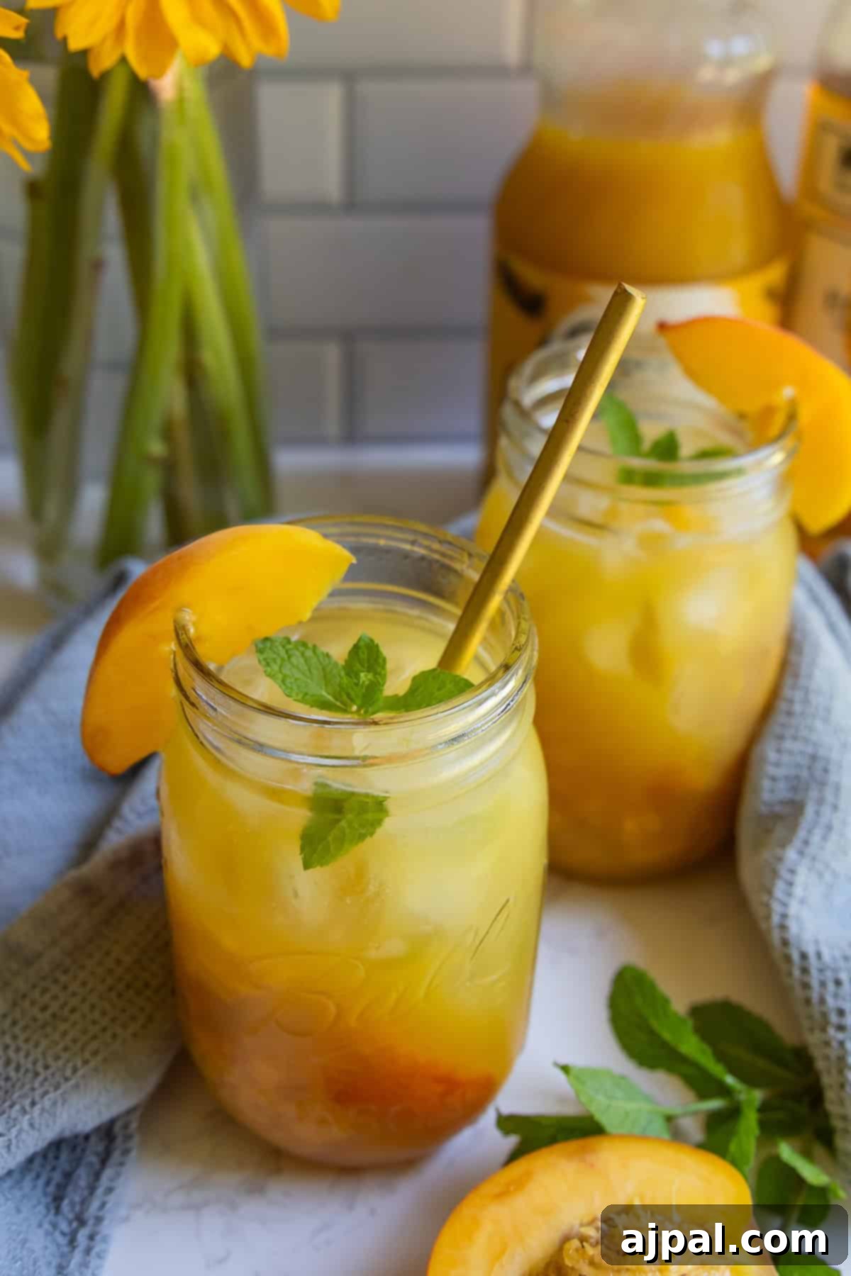Two mason jars filled with iced peach tea, garnished with mint leaves and peach slices, sit on a table near a blue cloth and fresh peaches—perfect for inspiring a refreshing bourbon peach smash cocktail.