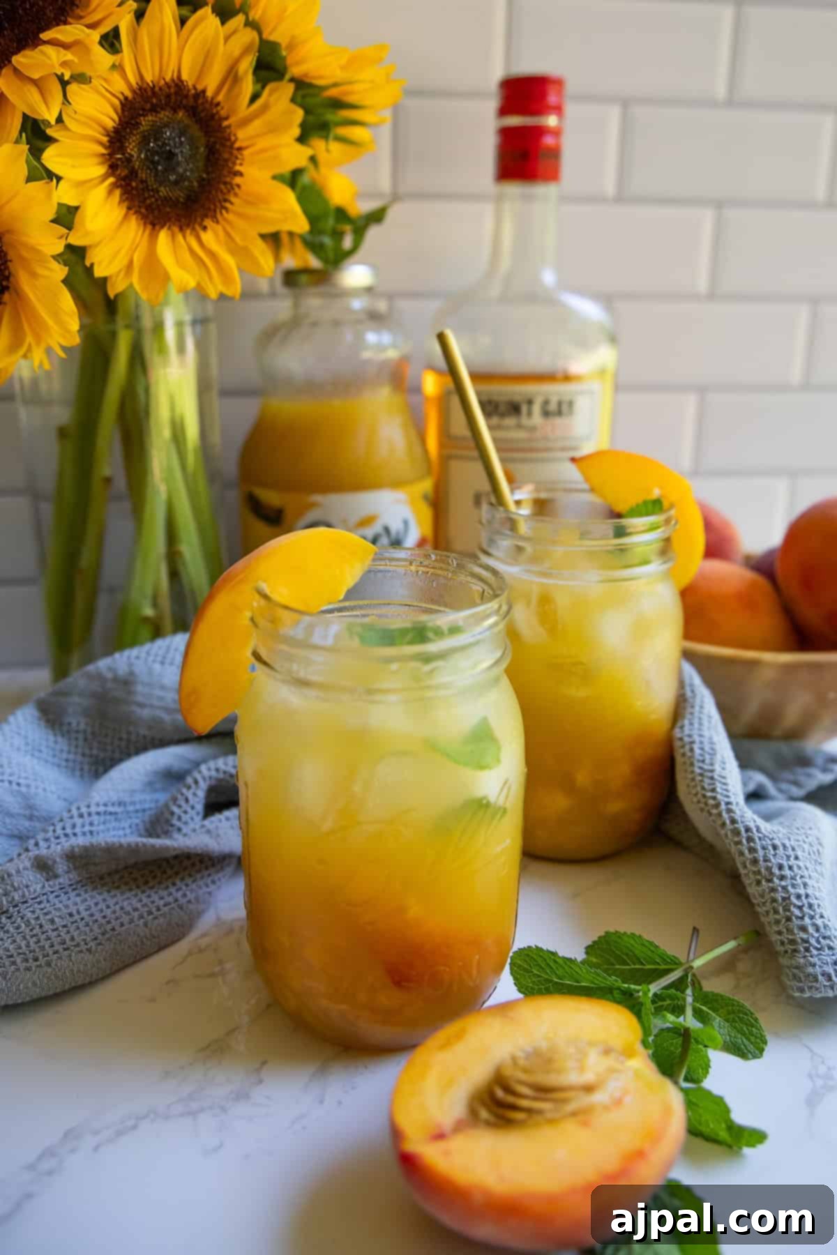 Two mason jars filled with a bourbon peach smash cocktail, garnished with peach slices and mint, sit on a countertop with a bottle of rum, peaches, and sunflowers in the background.