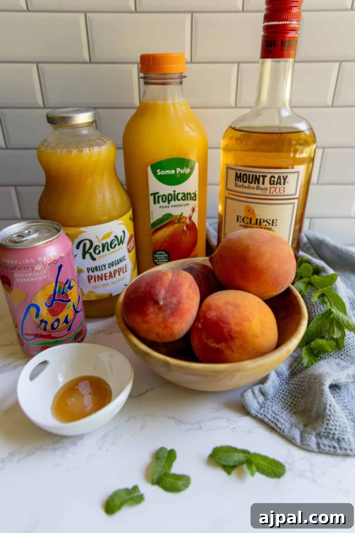 A wooden bowl of peaches sits on a counter with bottles of orange juice, pineapple juice, rum, sparkling water, fresh mint, and honey—perfect ingredients for crafting a refreshing bourbon peach smash cocktail.