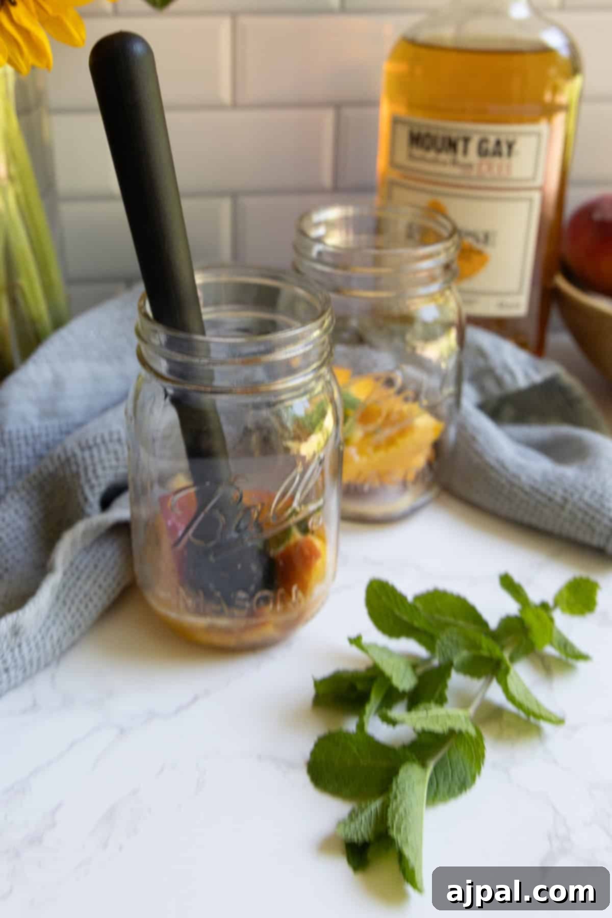 Two mason jars with muddled fruit for a bourbon peach smash cocktail sit on a countertop, a muddler nearby. A bottle of Mount Gay rum is in the background, with mint leaves and a gray cloth completing the scene.