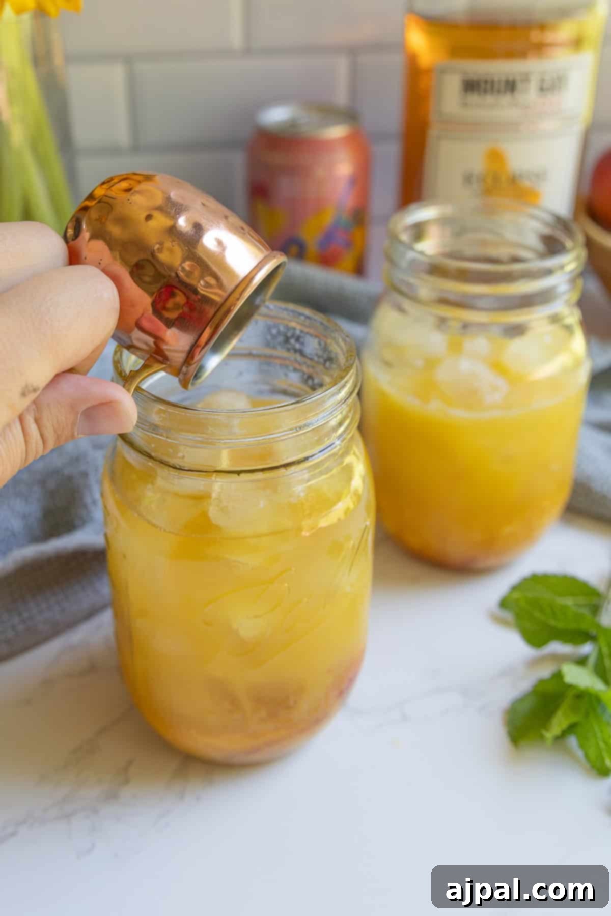 A hand pours liquid from a small copper cup into a mason jar filled with ice and an orange bourbon peach smash cocktail; another similar jar sits nearby on the marble counter.