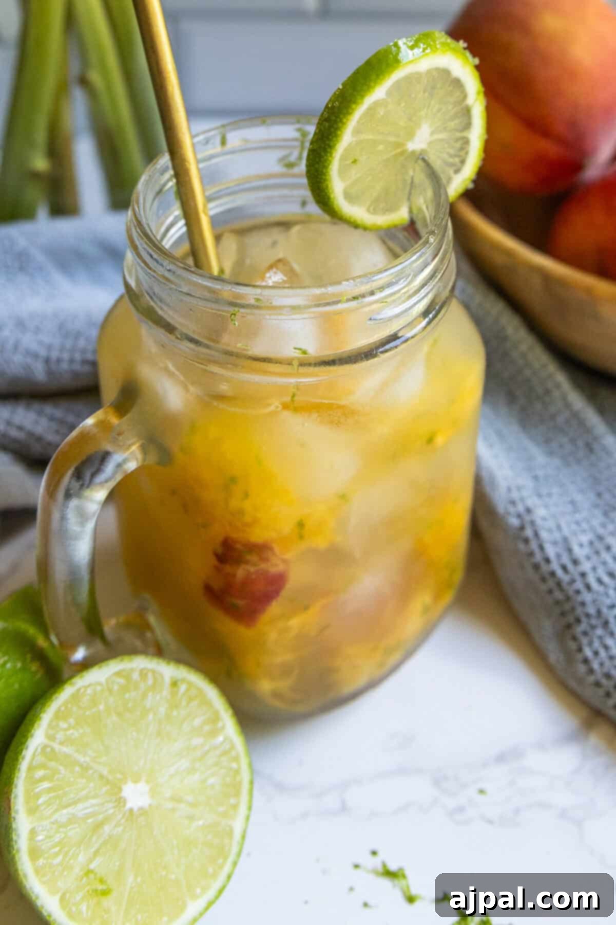 A mason jar filled with a peach La Croix mocktail, iced and garnished with a lime wheel, gold straw, and visible fruit pieces; fresh limes and peaches are nearby.