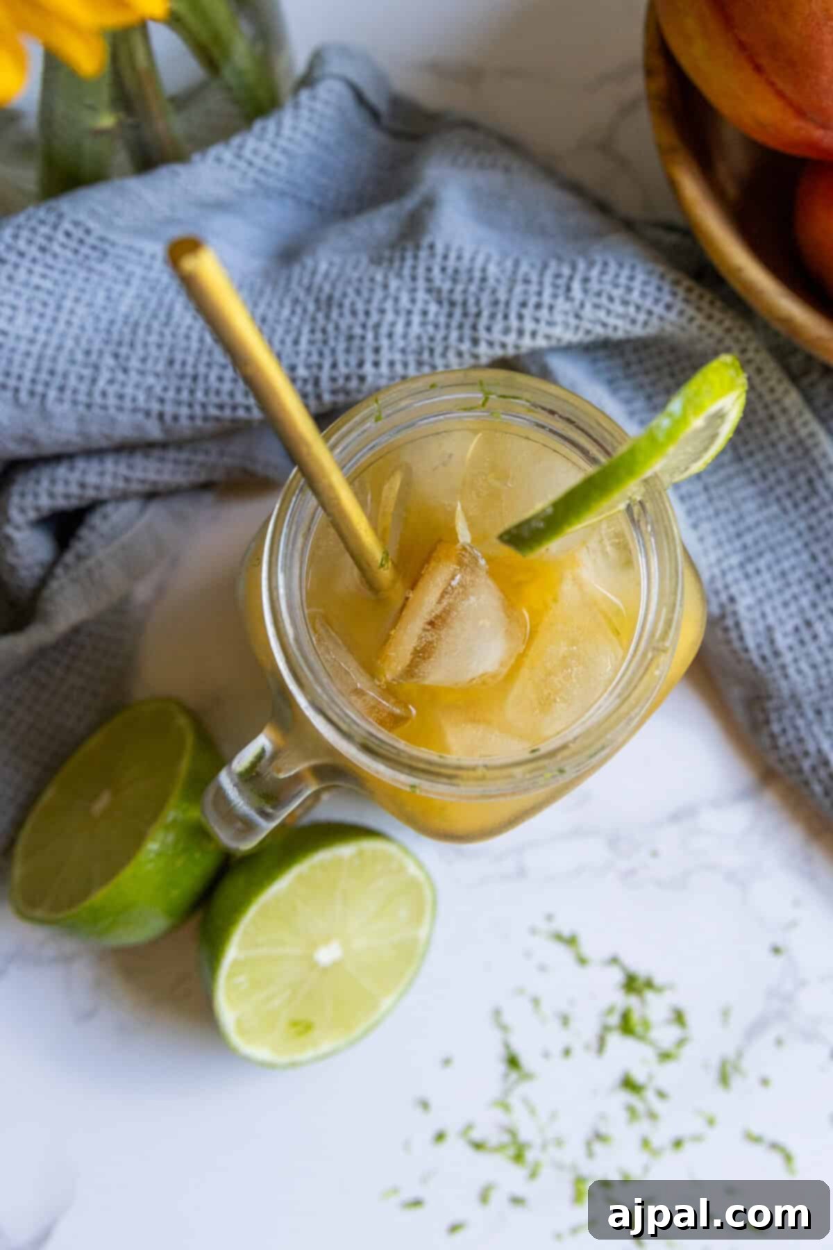A glass mug with a refreshing peach La Croix mocktail, iced and garnished with a lime wedge and gold straw, sits beside two lime halves on a white surface with a blue cloth.