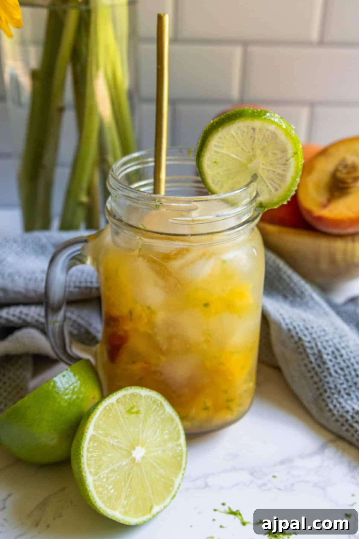 A mason jar filled with a peach La Croix mocktail, garnished with a lime slice on the rim, sits on a counter with a cut lime and a peach in the background.