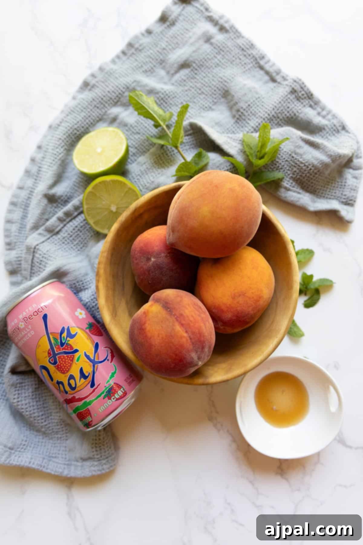 A wooden bowl with peaches, a can of La Croix, fresh mint, lime halves, and a small dish of honey are arranged on a cloth—perfect ingredients for crafting a refreshing peach La Croix mocktail.