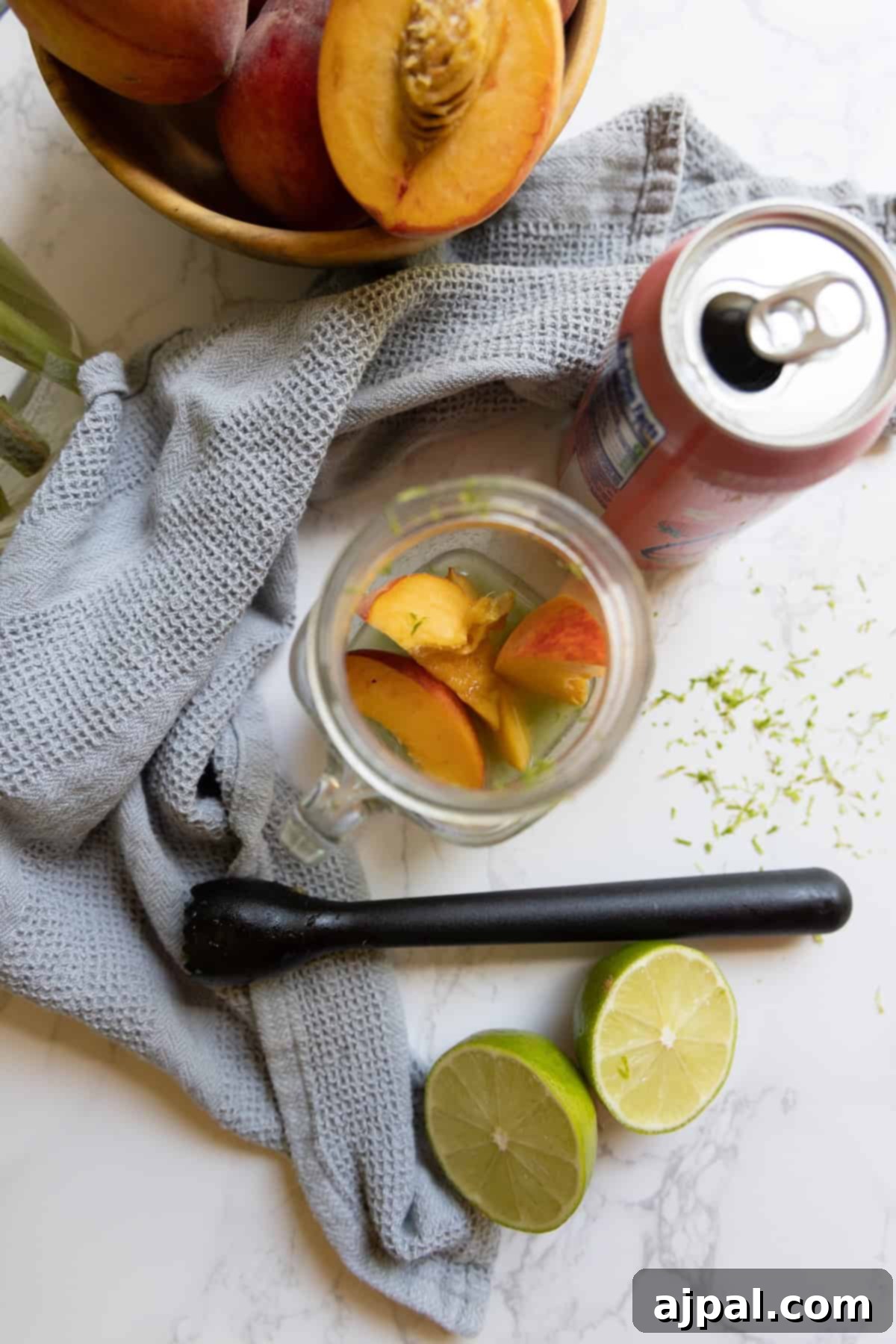 A mason jar with peach slices, a halved lime, a peach, a can of Peach La Croix mocktail, a black muddler, a gray cloth, and a bowl of peaches rest on a marble surface.