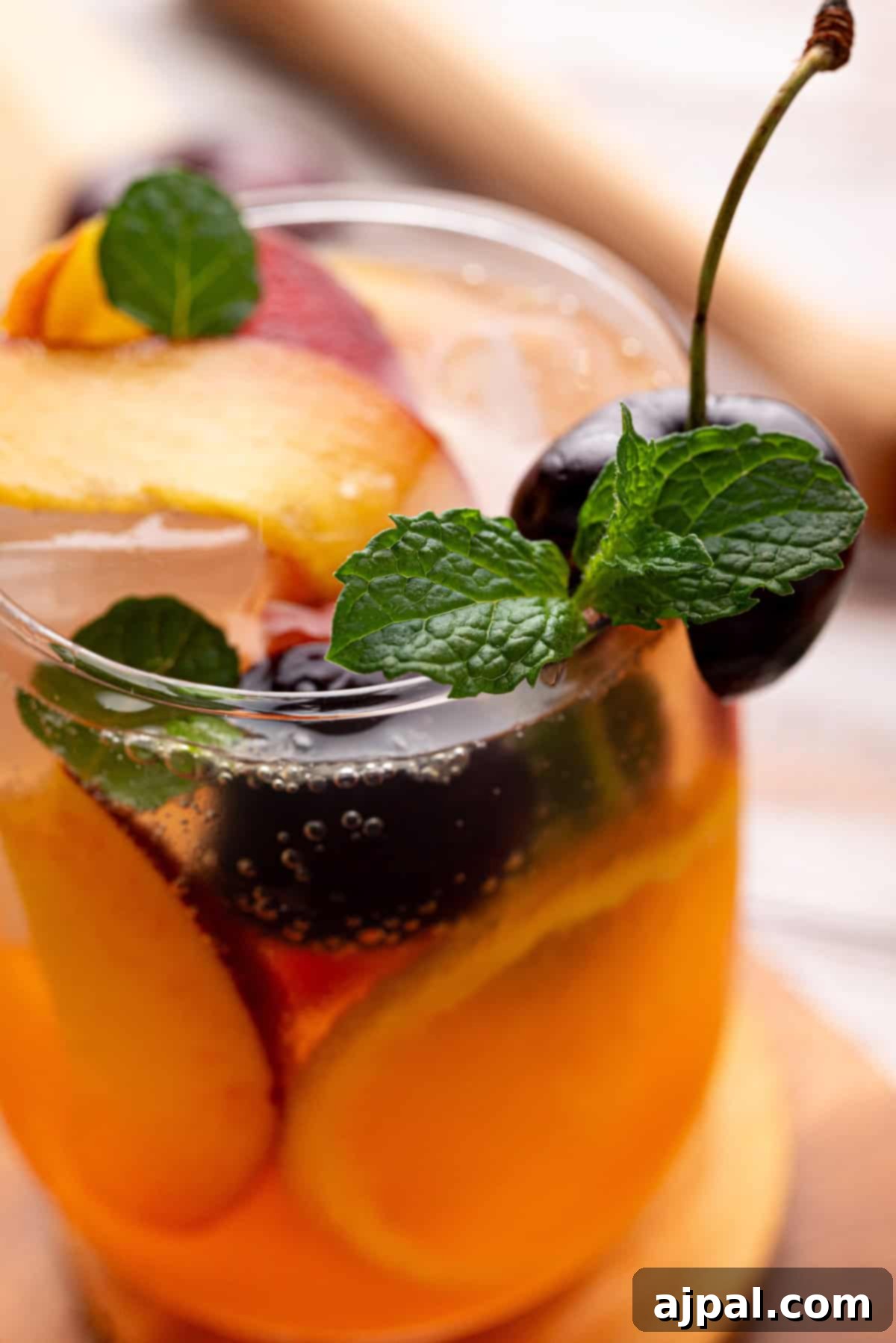 Vibrant Stone Fruit Refresher 2 A close-up of a glass filled with an orange-colored Sangria Mocktail, ice, stone fruit slices like peach, cherries, and a sprig of mint.