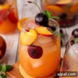 A glass of iced stone fruit mocktail featuring peach and cherry, garnished with fresh fruit slices and mint, sits on a wooden surface.