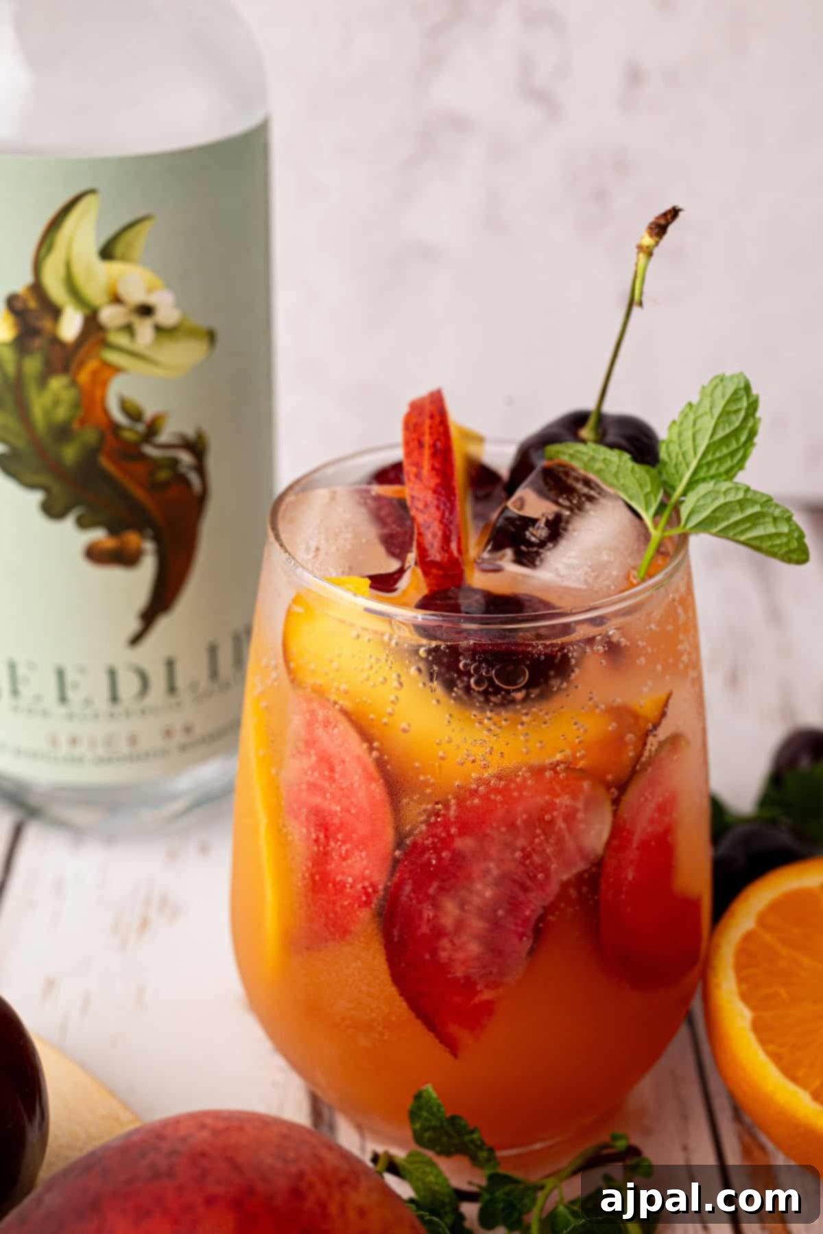 Vibrant Stone Fruit Refresher 4 A glass of iced fruit cocktail mocktail with peach slices, cherries, and mint garnish sits beside a bottle labeled "Seedlip" and fresh stone fruit on a wooden surface.