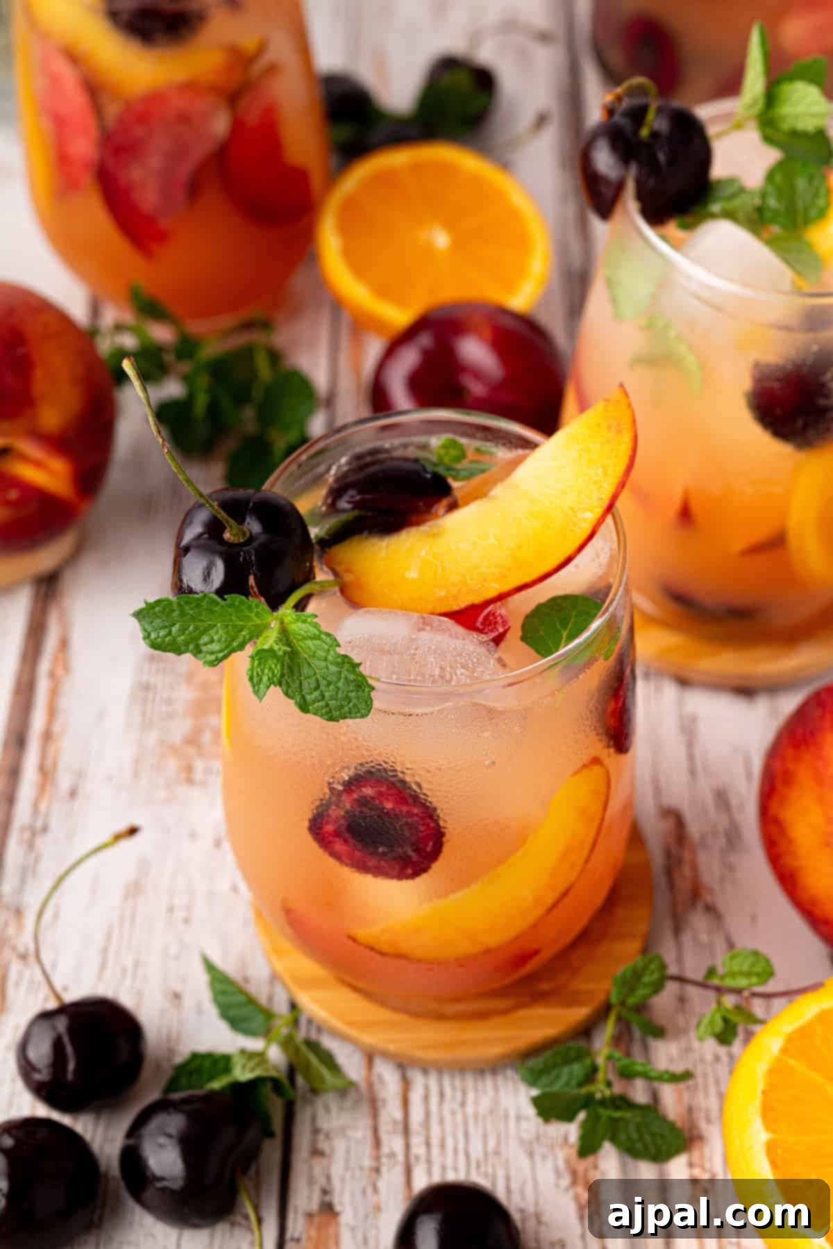 Vibrant Stone Fruit Refresher 5 A glass of iced sangria mocktail garnished with peach slices, cherries, and mint, surrounded by fresh stone fruit and mint on a wooden surface.