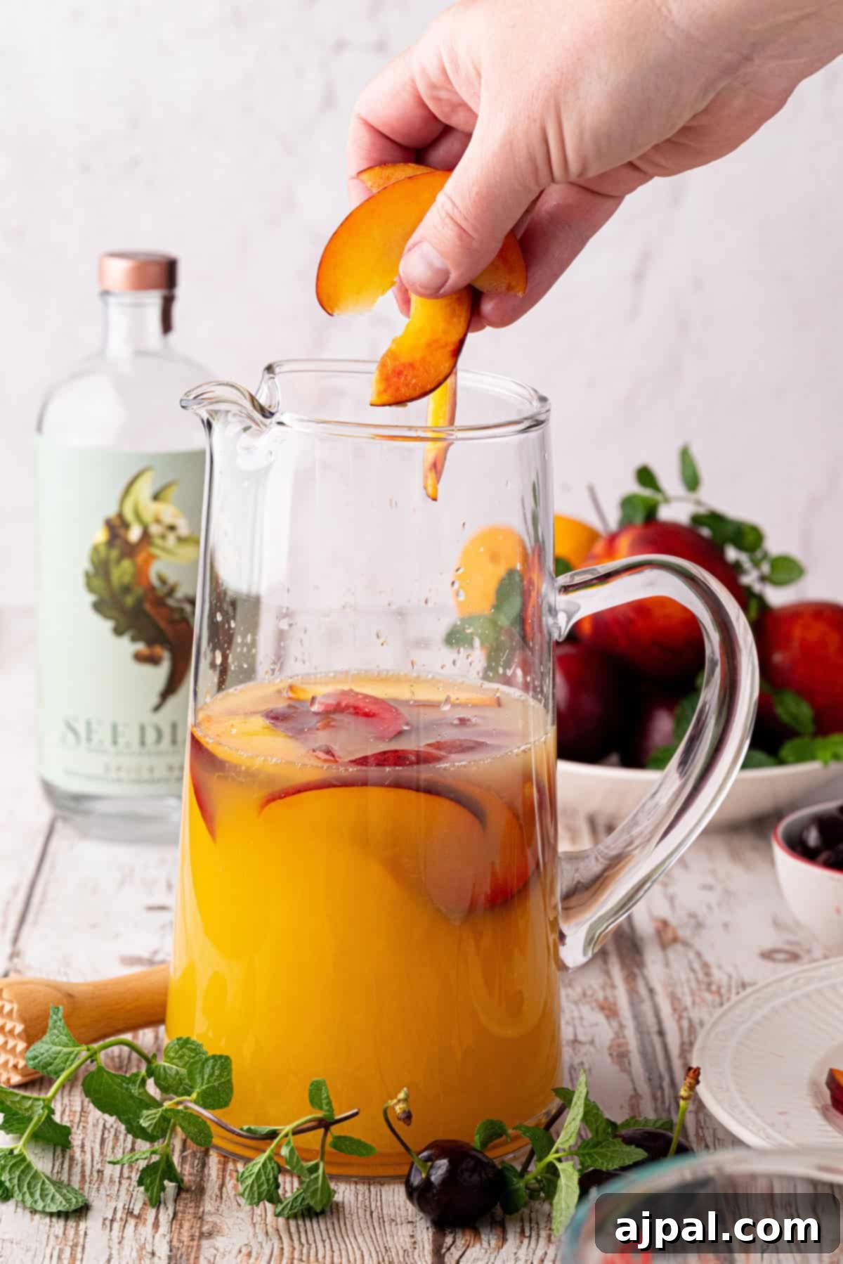 Vibrant Stone Fruit Refresher 7 A hand places a juicy stone fruit slice into a glass pitcher filled with yellow-orange sangria mocktail and fresh fruit slices, with a bottle, mint, and more fruit in the background.
