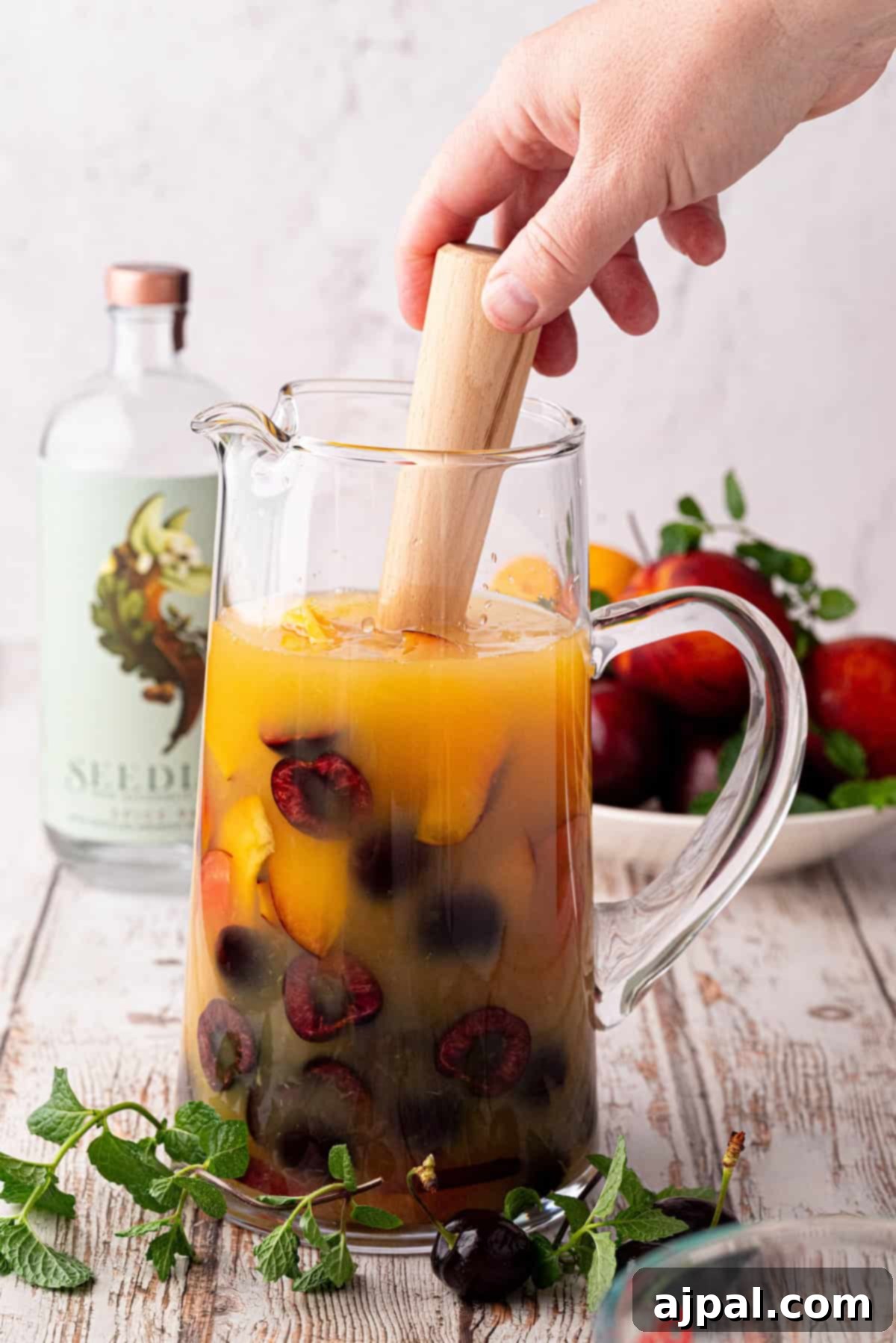 Vibrant Stone Fruit Refresher 8 A hand uses a wooden muddler to mix stone fruit and juice in a glass pitcher, crafting a refreshing sangria; a bottle and a plate of fruit are visible in the background.