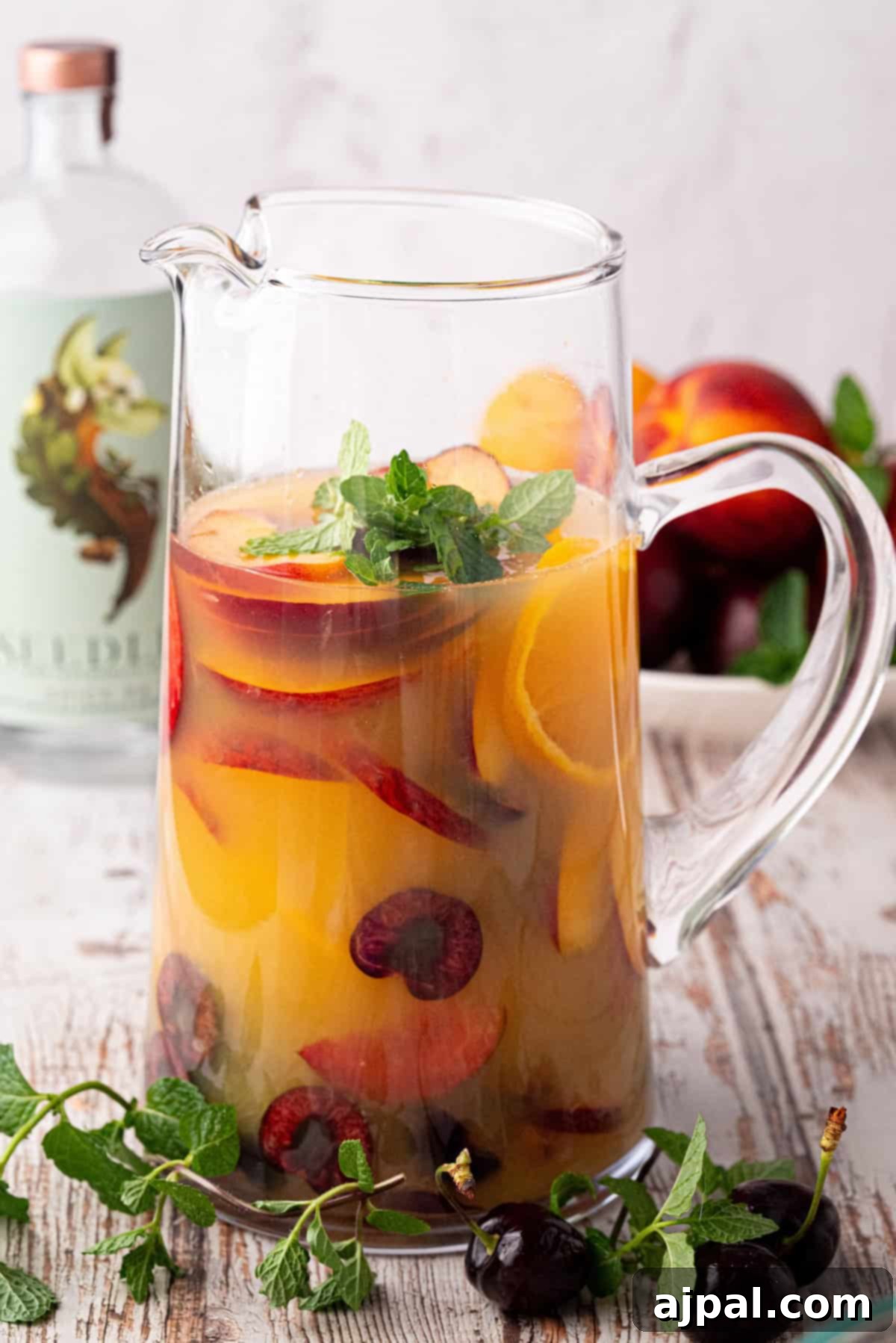 Vibrant Stone Fruit Refresher 9 A glass pitcher filled with a refreshing sangria mocktail featuring orange slices, cherries, and mint, with a bottle and bowl of stone fruit in the background.