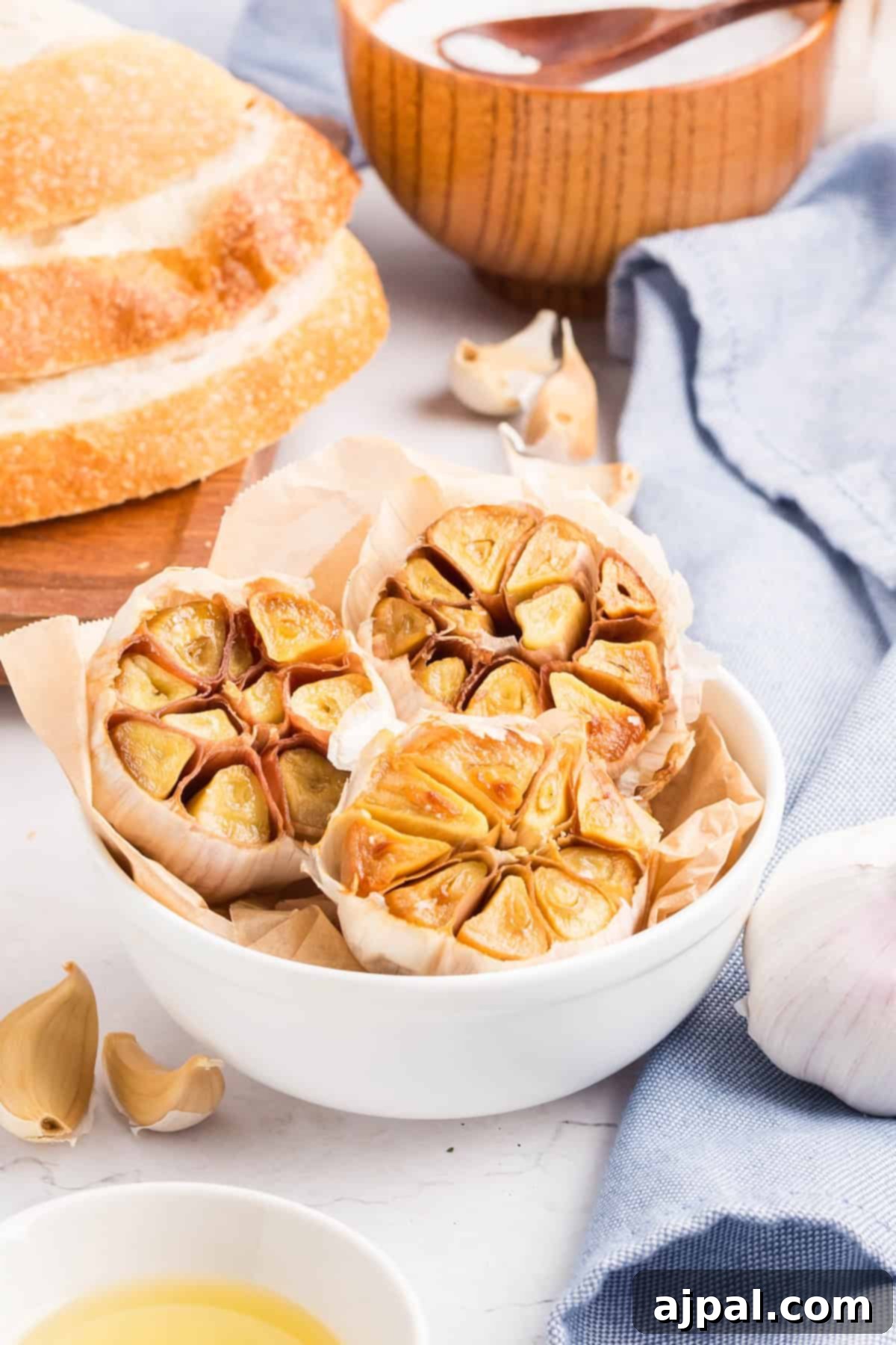 A white bowl filled with roasted garlic bulbs on parchment paper, surrounded by bread, a wooden bowl, linen, and scattered roasted garlic cloves.