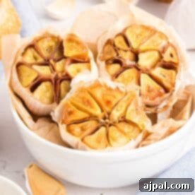 A white bowl filled with roasted garlic—halved heads glistening on parchment paper—sits amid raw garlic cloves on a light surface.