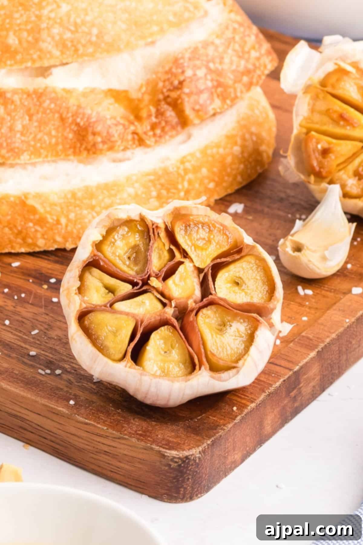A head of roasted garlic sits on a wooden cutting board near sliced bread and raw garlic cloves, with coarse salt scattered around, highlighting the rich aroma of fresh roasted garlic.