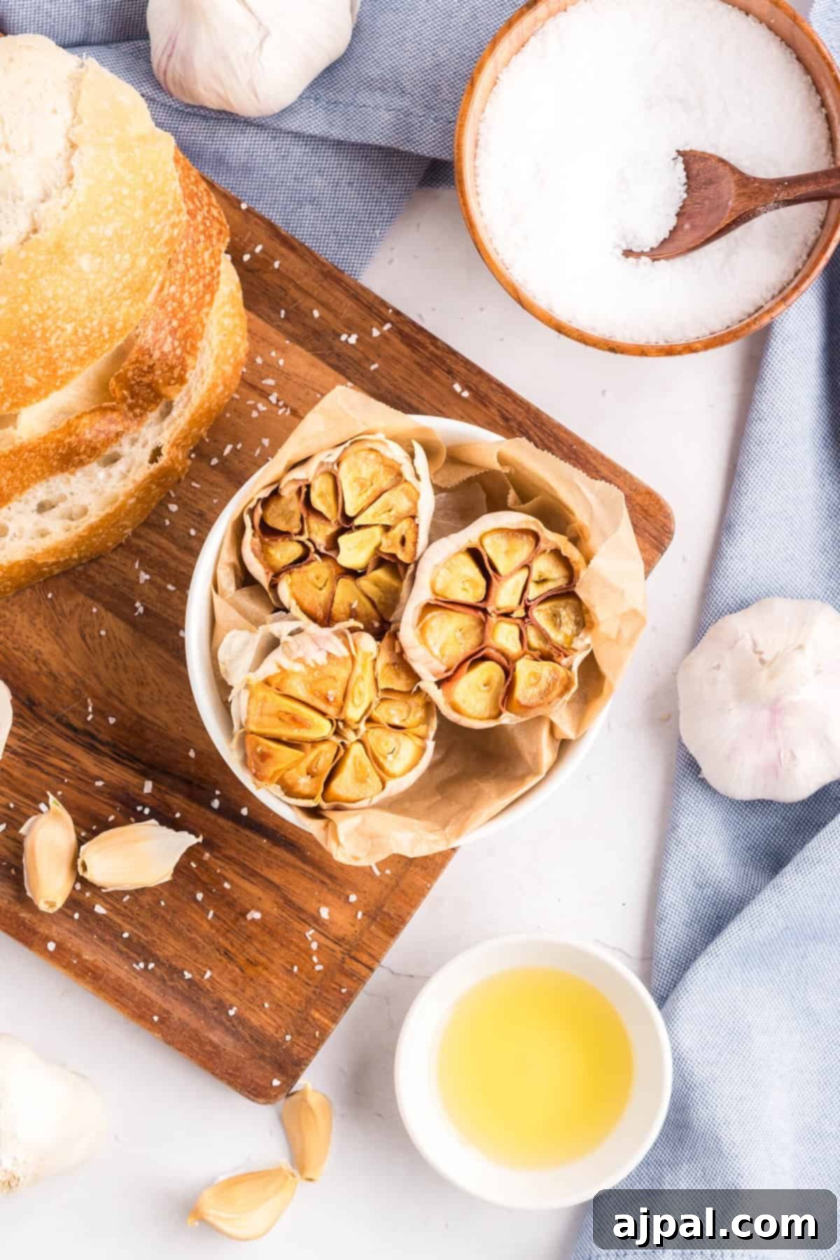 A bowl of roasted garlic heads on parchment paper, next to sliced bread, a wooden bowl of salt, a small bowl of oil, and fresh garlic cloves on a wooden board—perfect for enjoying the rich flavor of roasted garlic.