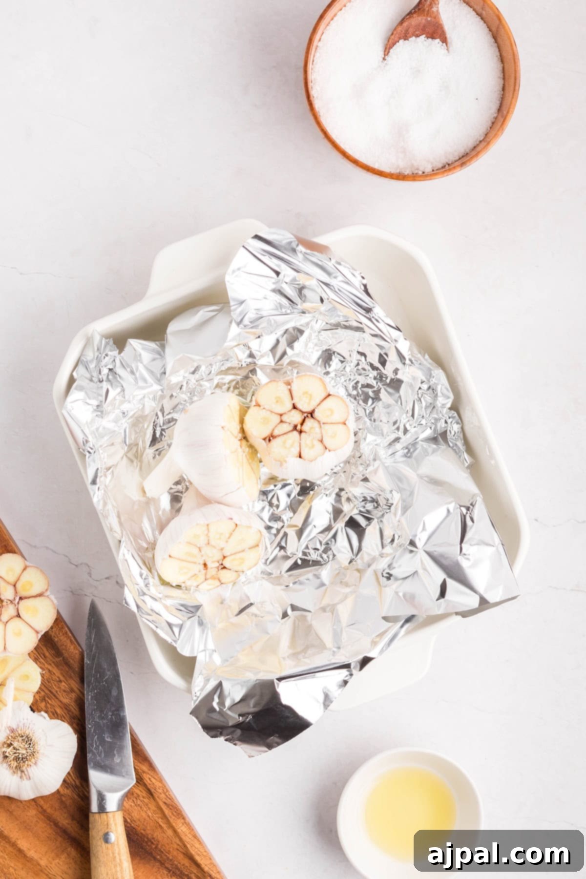 A baking dish with halved garlic bulbs ready for roasted garlic rests on aluminum foil, alongside a knife, a bowl of salt, and a small bowl of oil, all arranged on a white surface.