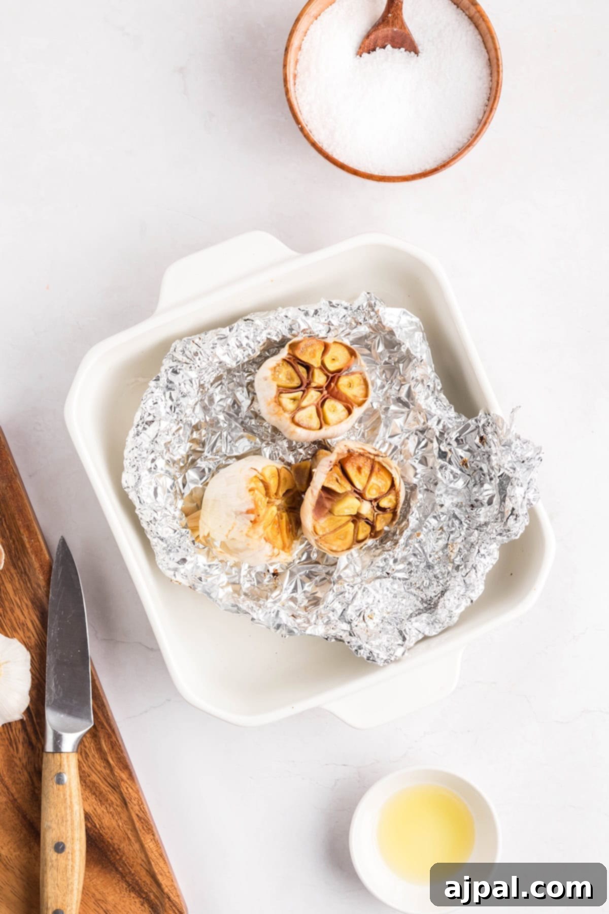 A head of roasted garlic cut in half sits on aluminum foil in a white baking dish, with a knife, a bowl of salt, and a small cup of oil nearby—perfect for spreading this flavorful roasted garlic on your favorite dishes.