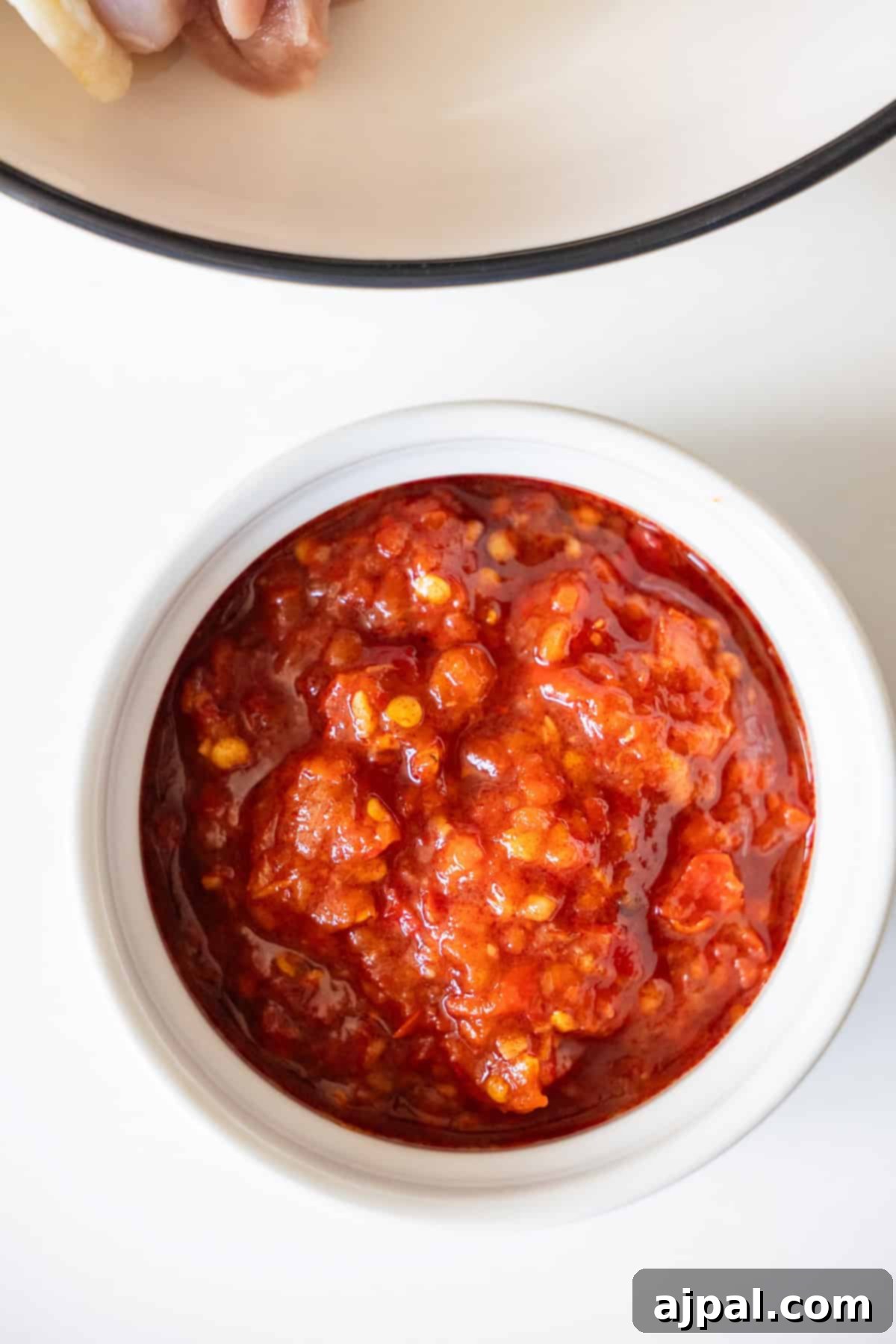 Zesty Harissa Chicken Thighs with Briny Olives 6 A white bowl filled with chunky red chili paste, perfect for serving alongside spicy chicken thighs, is placed on a white surface.