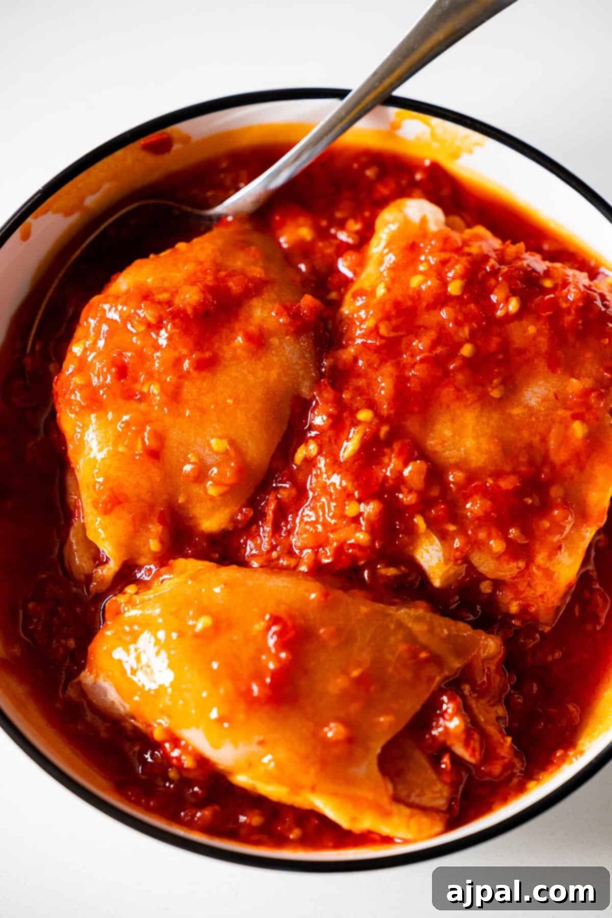 Zesty Harissa Chicken Thighs with Briny Olives 7 A bowl with three pieces of spicy chicken covered in a thick, red chili sauce, served with smashed olives and a spoon resting on the side.