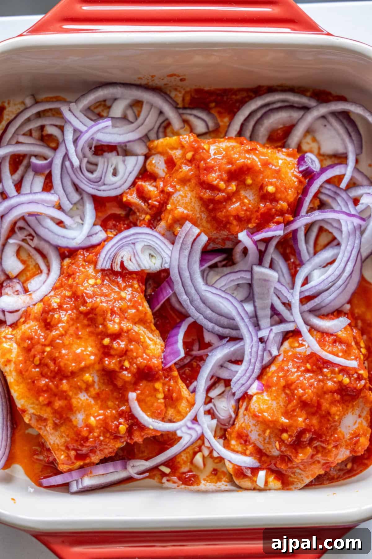 Zesty Harissa Chicken Thighs with Briny Olives 8 Raw chicken pieces coated in red sauce and topped with sliced red onions in a rectangular baking dish—perfect for making flavorful Harissa Chicken Thighs.