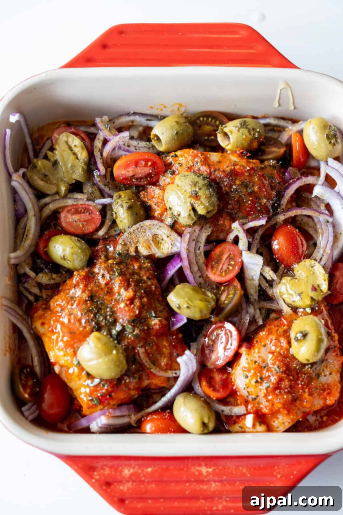 Zesty Harissa Chicken Thighs with Briny Olives 10 Baking dish with harissa chicken thighs, halved cherry tomatoes, sliced red onions, and smashed olives, ready to be cooked.