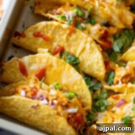Baked Chicken Tacos with a crispy hard shell are filled with shredded chicken, melted cheese, chopped onions, cilantro, and drizzled with red sauce, all arranged closely together on a tray.