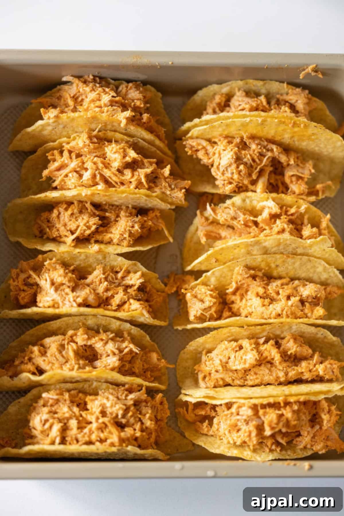 A baking tray holds rows of Baked Chicken Tacos, each hard shell filled with shredded, seasoned chicken, arranged neatly for baking or serving.