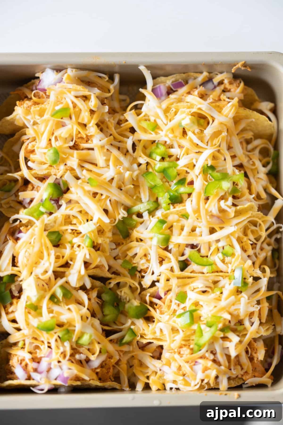A baking dish filled with tortilla chips, shredded cheese, chopped green bell peppers, and red onions—just like the perfect Baked Chicken Tacos, ready to be baked to melty perfection.